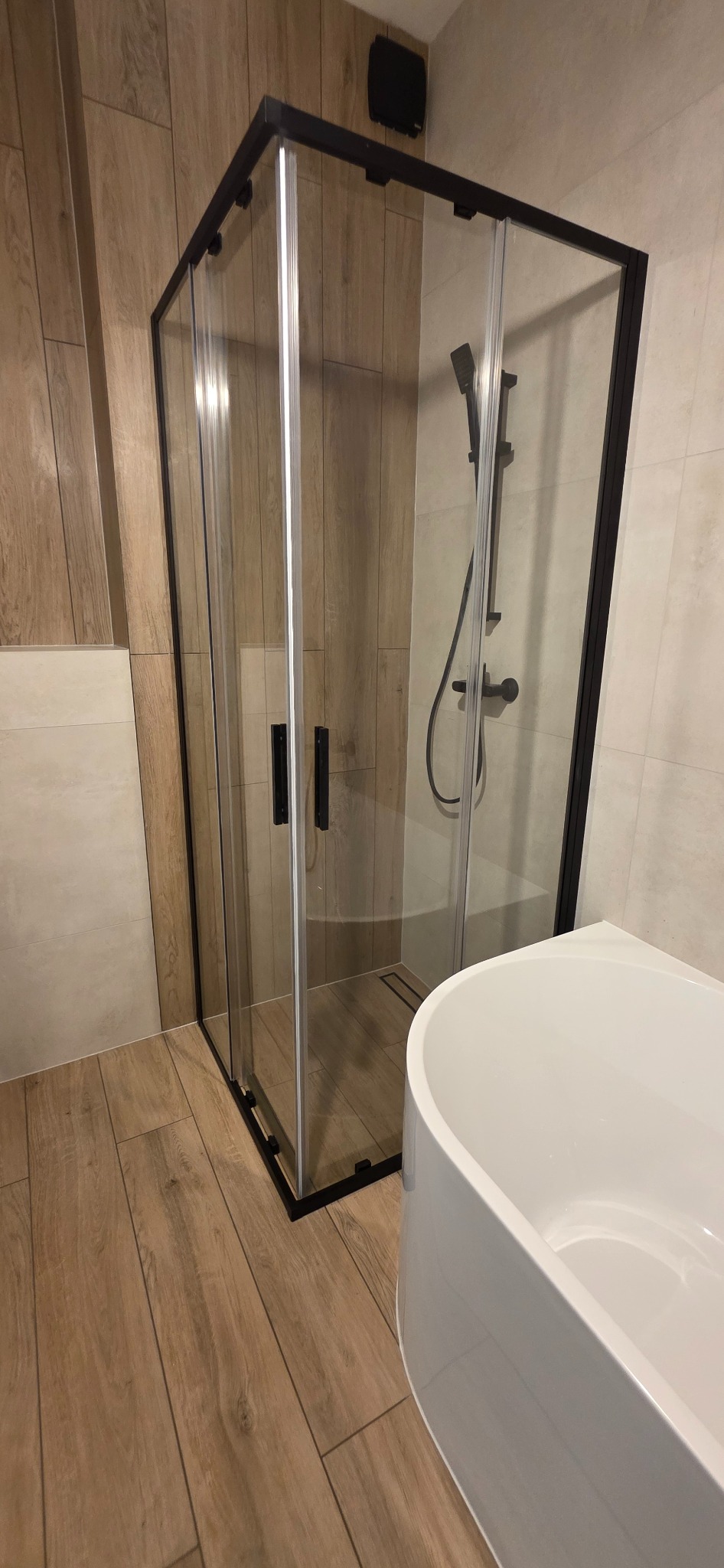 Nowoczesna kabina prysznicowa z czarnymi profilami i biała wanna w wykończonej łazience z płytkami imitującymi drewno na podłodze i ścianach.