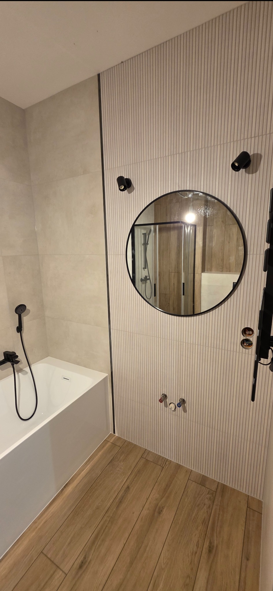 Nowoczesna łazienka z wanną, okrągłym lustrem i drewnopodobną podłogą. Ściany wyłożone płytkami z subtelnym, pionowym wzorem. Minimalistyczne oświetlenie punktowe.