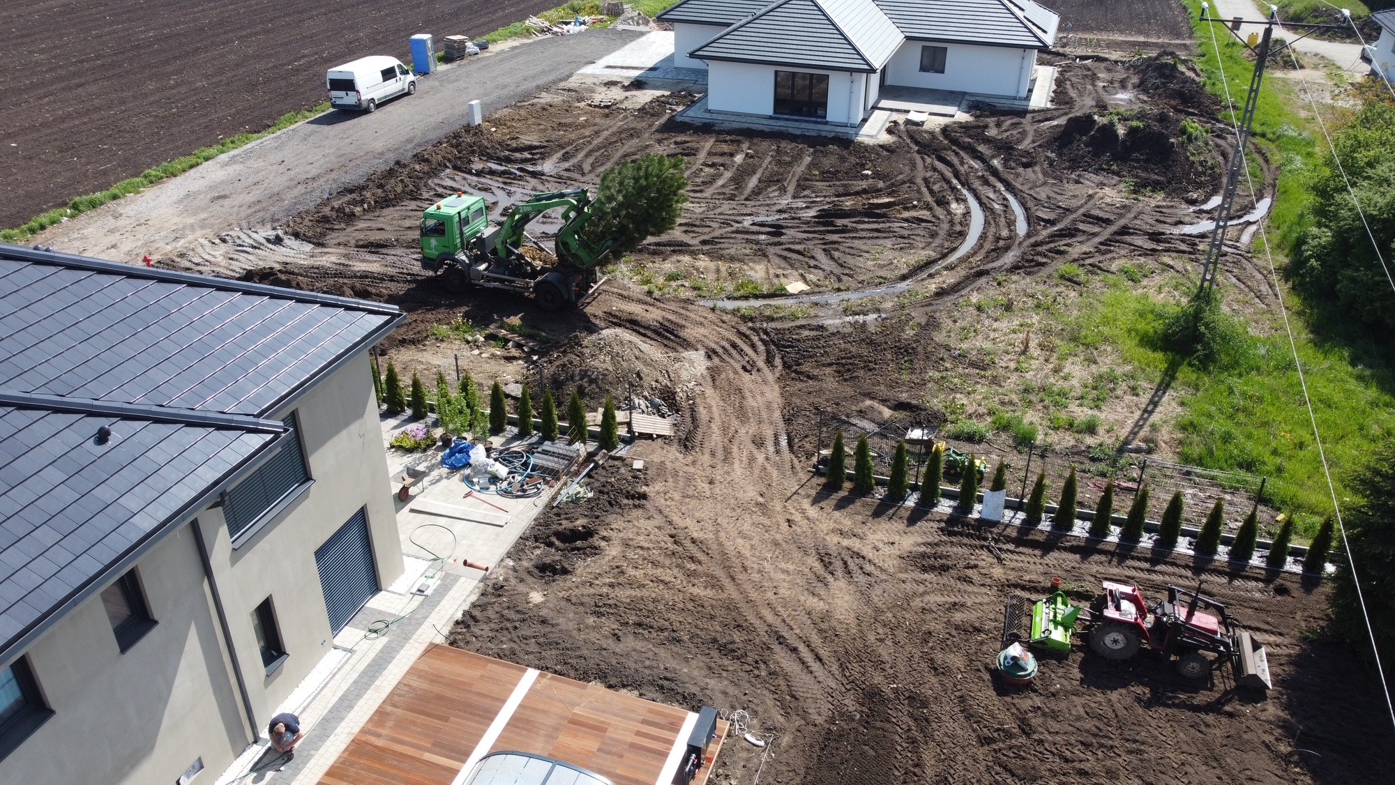 Widok z lotu ptaka na plac budowy z zielonym traktorem przesadzającym drzewo i czerwonym traktorem wyrównującym teren przy nowym domu.