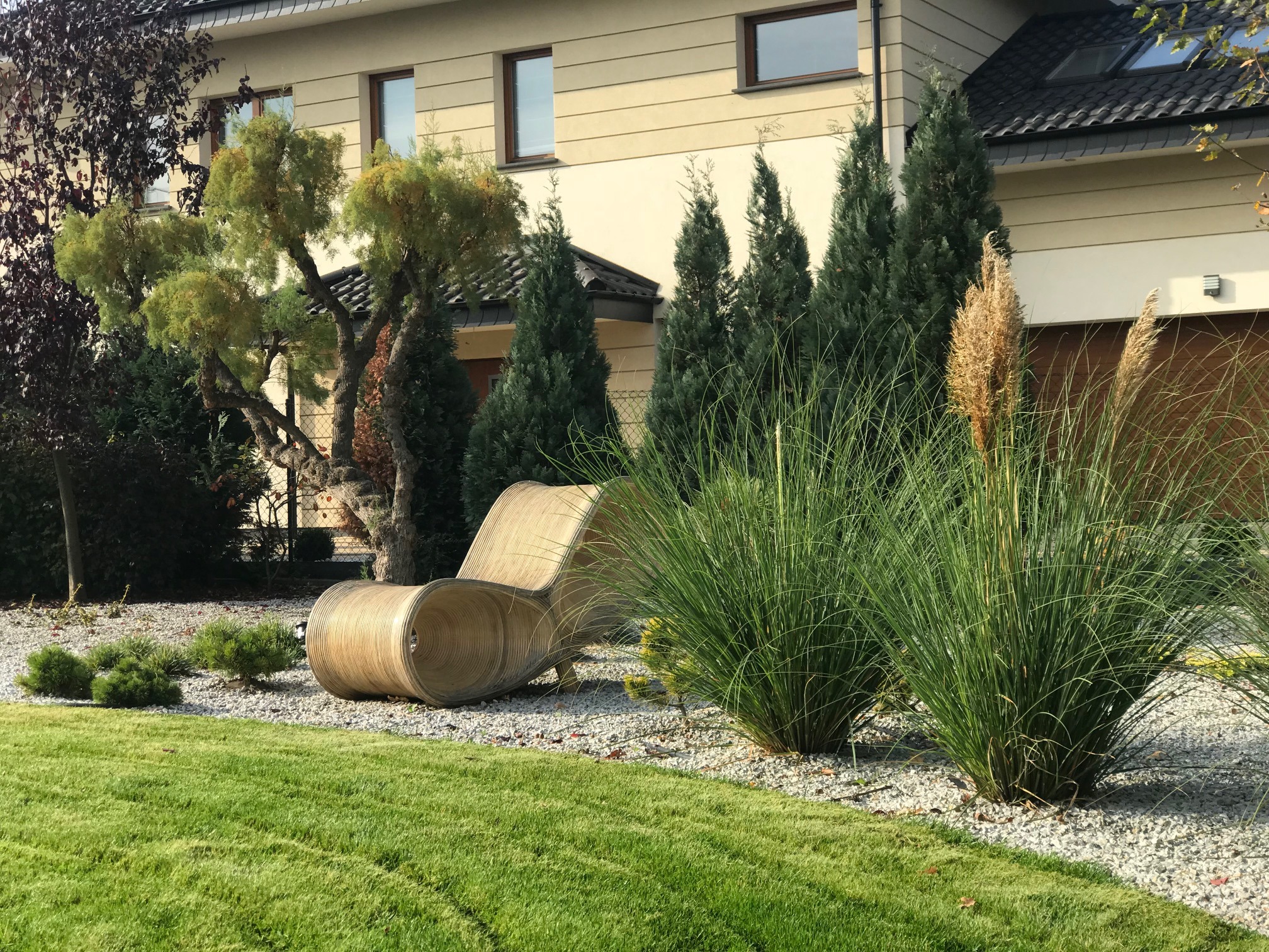 Nowoczesny ogród przydomowy z ozdobnymi trawami, drzewami iglastymi i oryginalnym, drewnianym siedziskiem. Białe kamyczki i trawnik dopełniają aranżację.