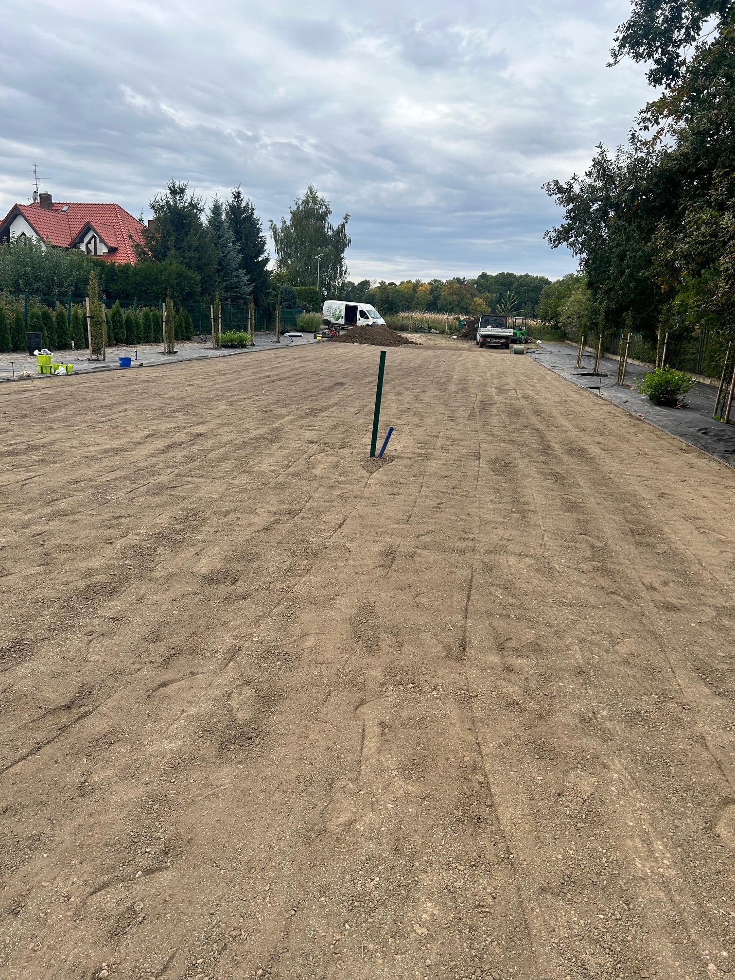 Przygotowany teren pod trawnik z widocznymi śladami po maszynach, w tle posadzone drzewa i krzewy. Na posesji stoją dwa samochody dostawcze.