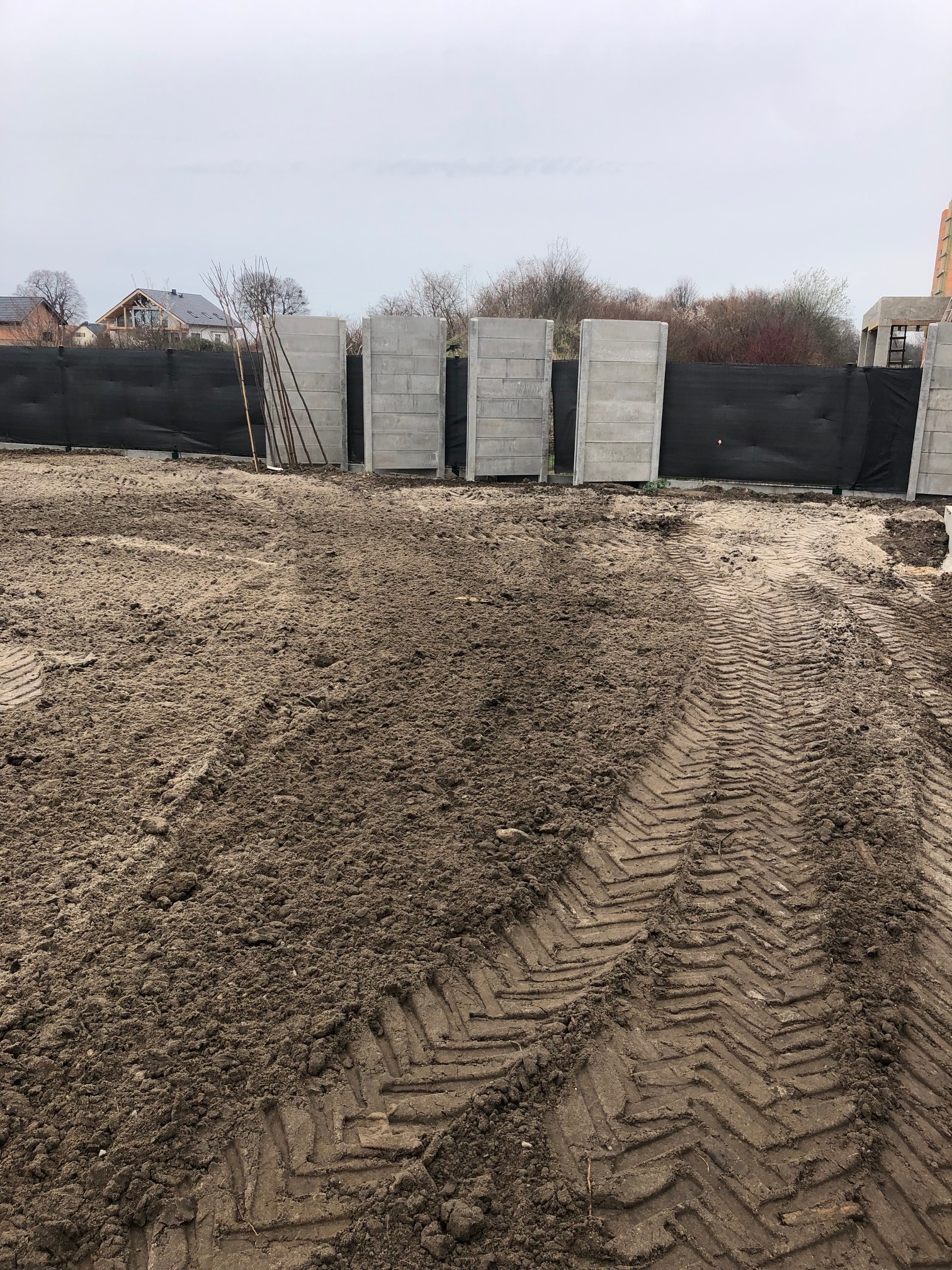 Przygotowany teren pod budowę z betonowymi prefabrykatami ogrodzeniowymi, widoczne ślady opon ciężkiego sprzętu i czarna siatka ogrodzeniowa.