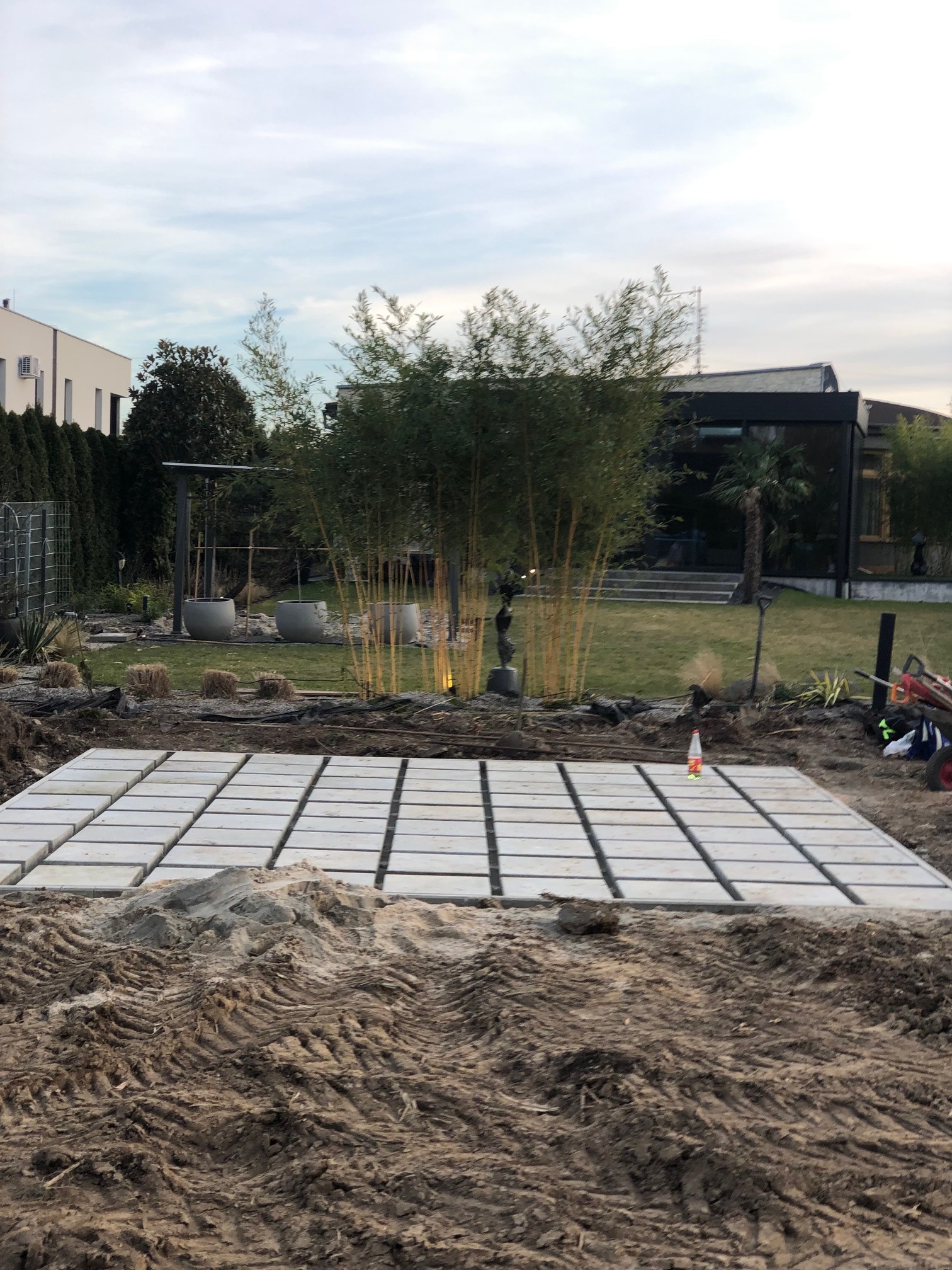 Układanie płyt chodnikowych w ogrodzie, widoczne przygotowane podłoże i częściowo ułożona nawierzchnia. W tle bambusy, trawnik i nowoczesny dom.