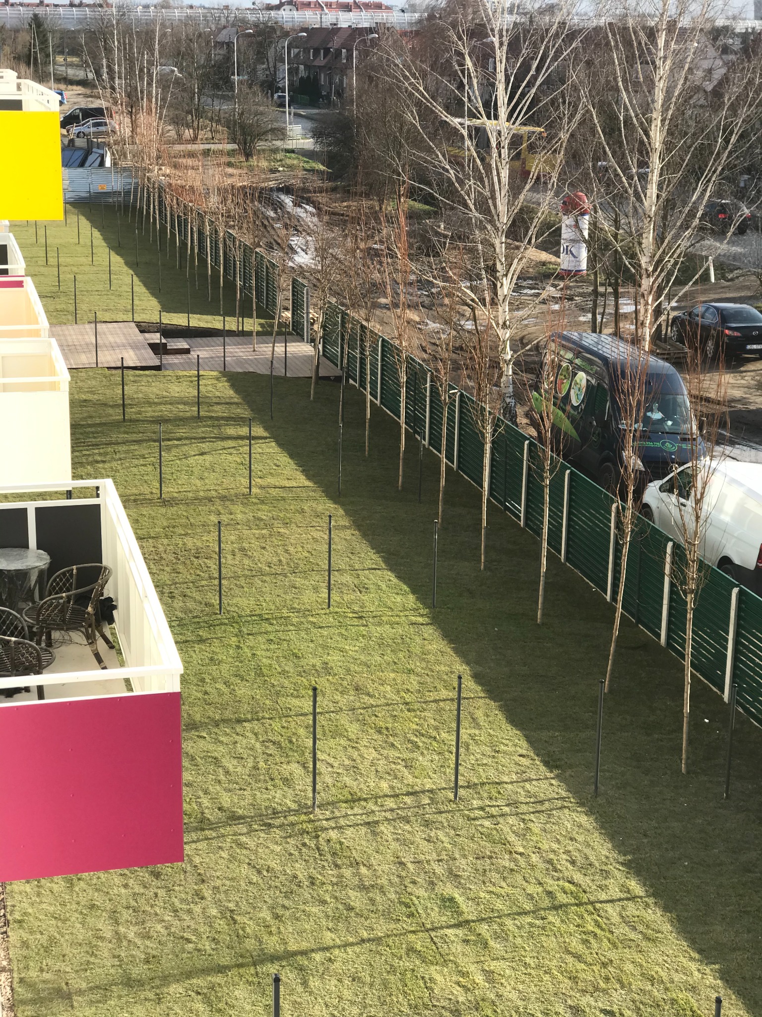 Widok z góry na założony trawnik z młodymi drzewkami wzdłuż ogrodzenia, z balkonami w lewym rogu i zaparkowanym busem z logo firmy ogrodniczej.