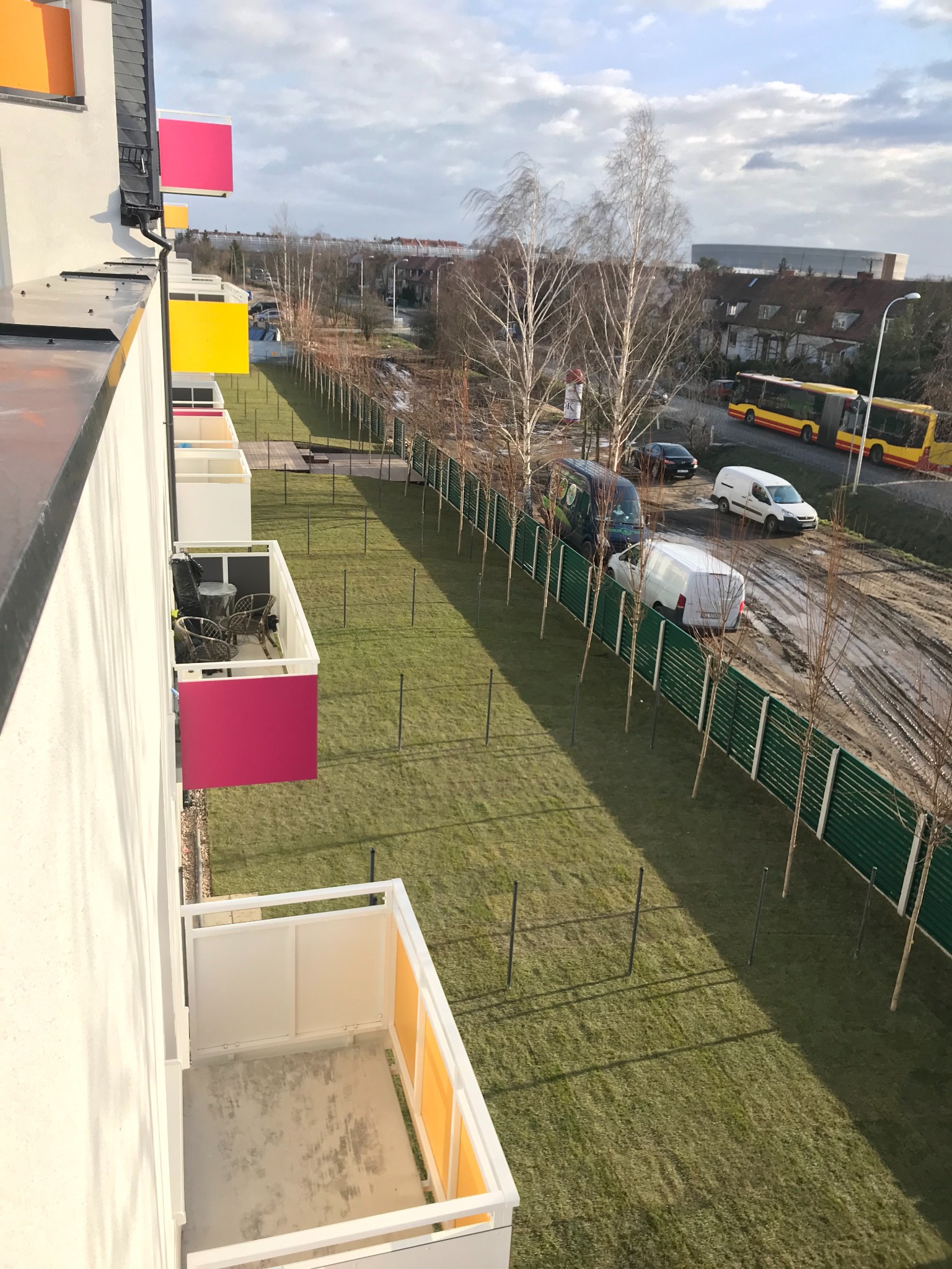 Widok z góry na nowoczesny budynek z kolorowymi balkonami, świeżo założonym trawnikiem i młodymi drzewami wzdłuż ogrodzenia. W tle ulica z pojazdami i budynki.