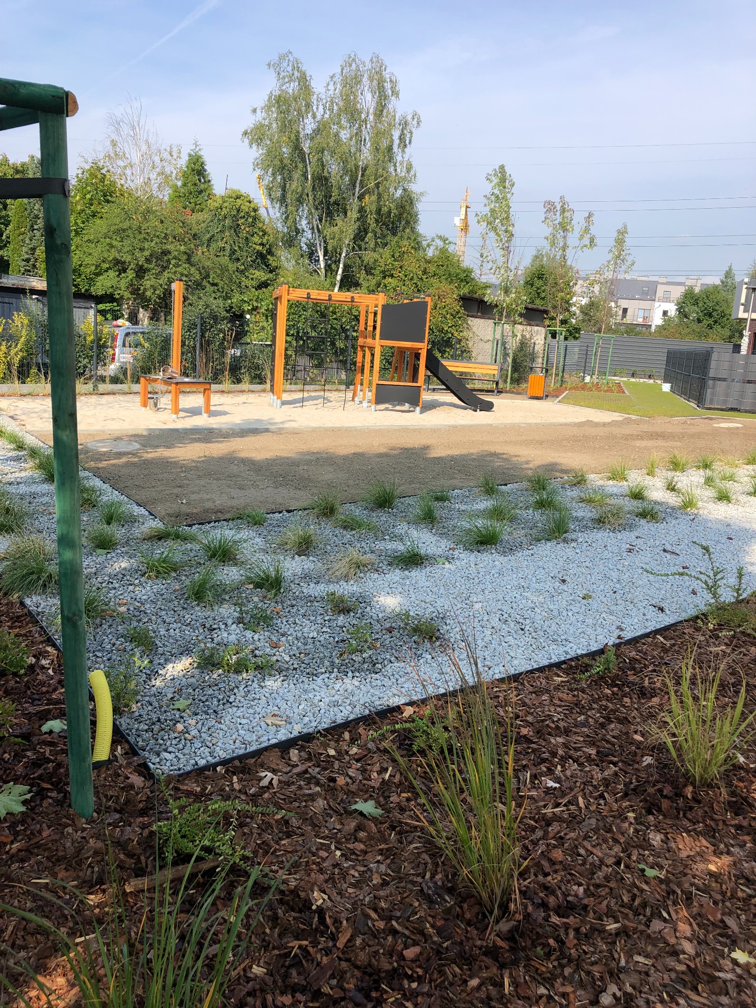 Nowoczesny ogród z placem zabaw: białe kamienie, trawa ozdobna, drewniane konstrukcje. W tle zieleń drzew i budynki. Kompozycja z perspektywą i dbałością o detale.