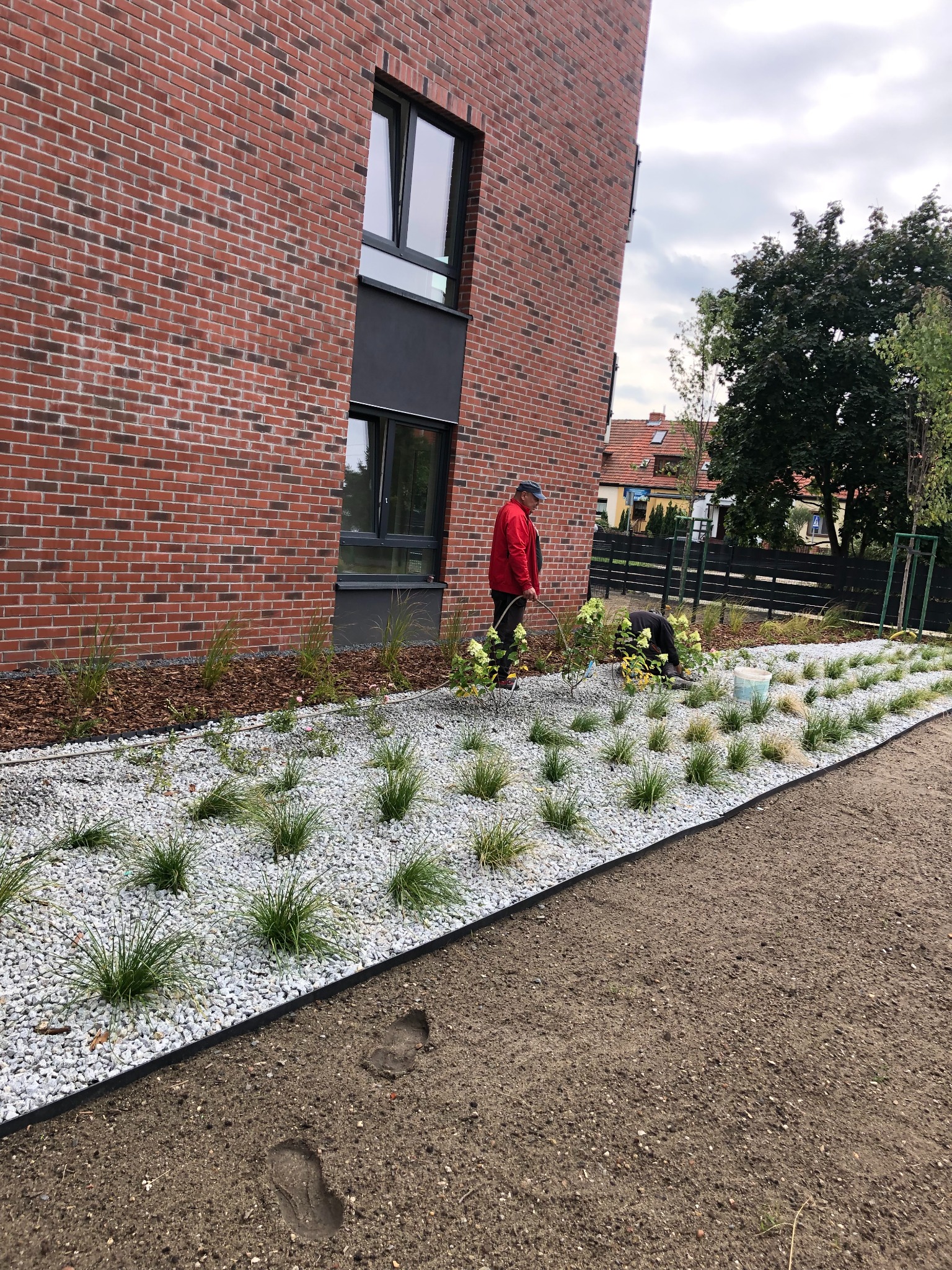 Ogród po założeniu: rabata z białym żwirem, trawami ozdobnymi i korą, na tle ceglanego budynku. Dwóch ogrodników podczas pracy.