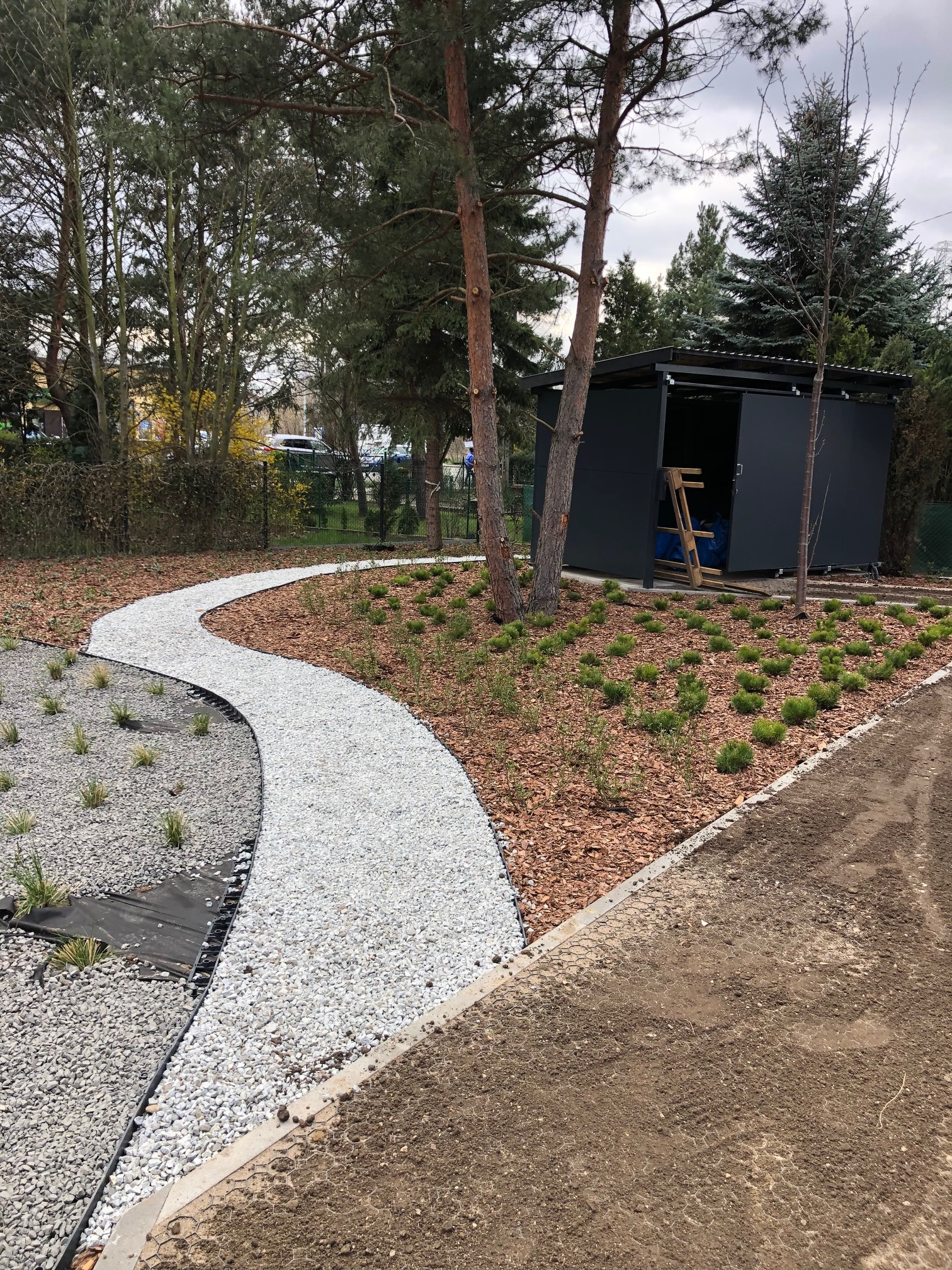 Ścieżka z białego żwiru wijąca się przez ogród z korą i młodymi roślinami, w tle szara szopa i drzewa iglaste. Widok na zagospodarowany teren.