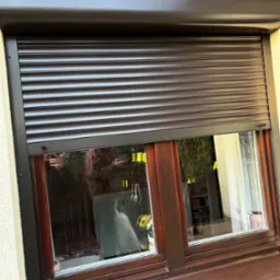 Ciemnobrązowa roleta na oknie z drewnianą ramą, częściowo opuszczona, widoczne odbicie osoby w oknie.