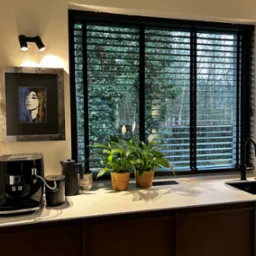 Czarne poziome osłony okienne w kuchni, z widokiem na zieleń za oknem, dwie rośliny doniczkowe na blacie, ekspres do kawy i czajnik obok obrazu w szarej ramie.