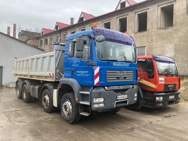 Dwa samochody ciężarowe MAN: niebieska wywrotka z napisem 'Holdschick' i czerwona z napisem 'Odpady', zaparkowane na betonowym placu, w tle budynek w stanie surowym.
