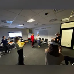 Grupa os&oacute;b w okularach VR uczestniczy w symulacji w sali szkoleniowej, z widocznym monitorem i napisem 'You say Jit, we say Team.' na ścianie.