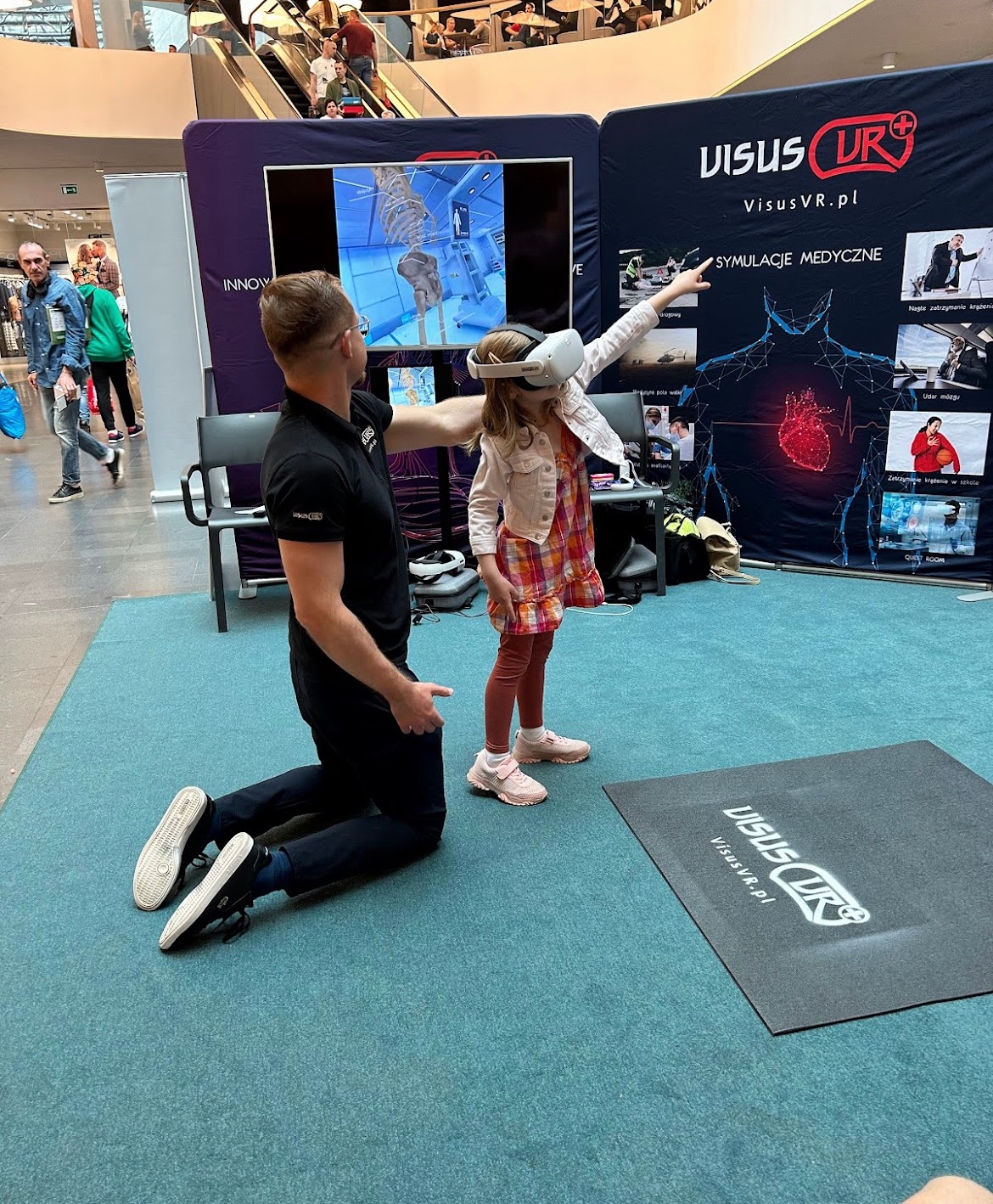 Dziewczynka w okularach VR uczestniczy w symulacji medycznej Visus VR, instruktor na kolanach pomaga jej, w tle ekran z modelem szkieletu i baner z logo Visus VR oraz symulacjami medycznymi.