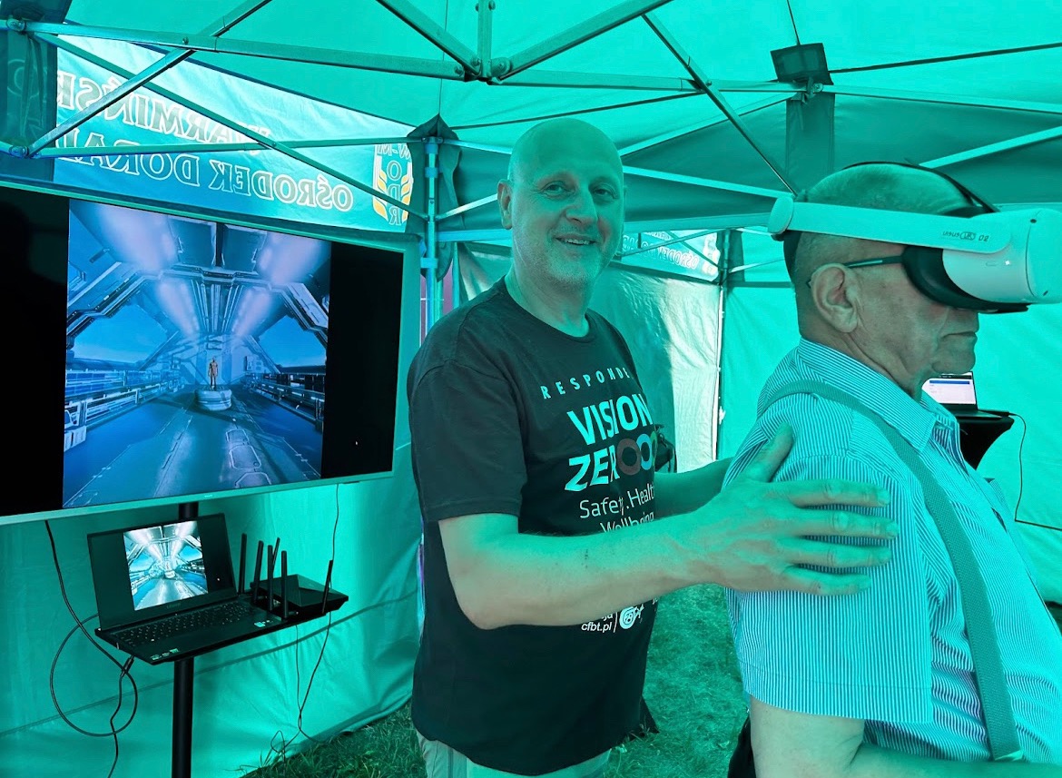 Mężczyzna z uśmiechem na twarzy, ubrany w koszulkę z napisem VISION ZERO, pomaga starszemu panu w okularach VR, w tle widoczny ekran z symulacją komputerową, prezentacja w namiocie.