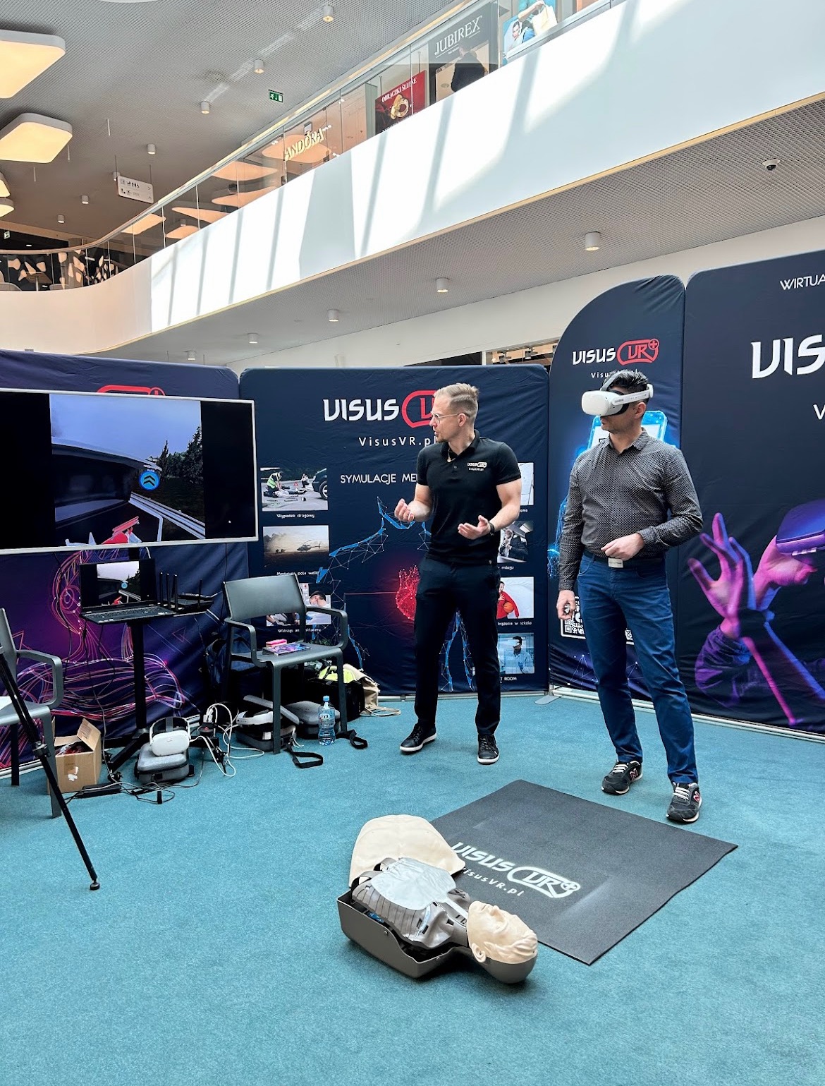 Prezentacja symulacji VR pierwszej pomocy w centrum handlowym, manekin do resuscytacji na podłodze, uczestnik w goglach VR, instruktor obok ekranu z widokiem z symulacji.