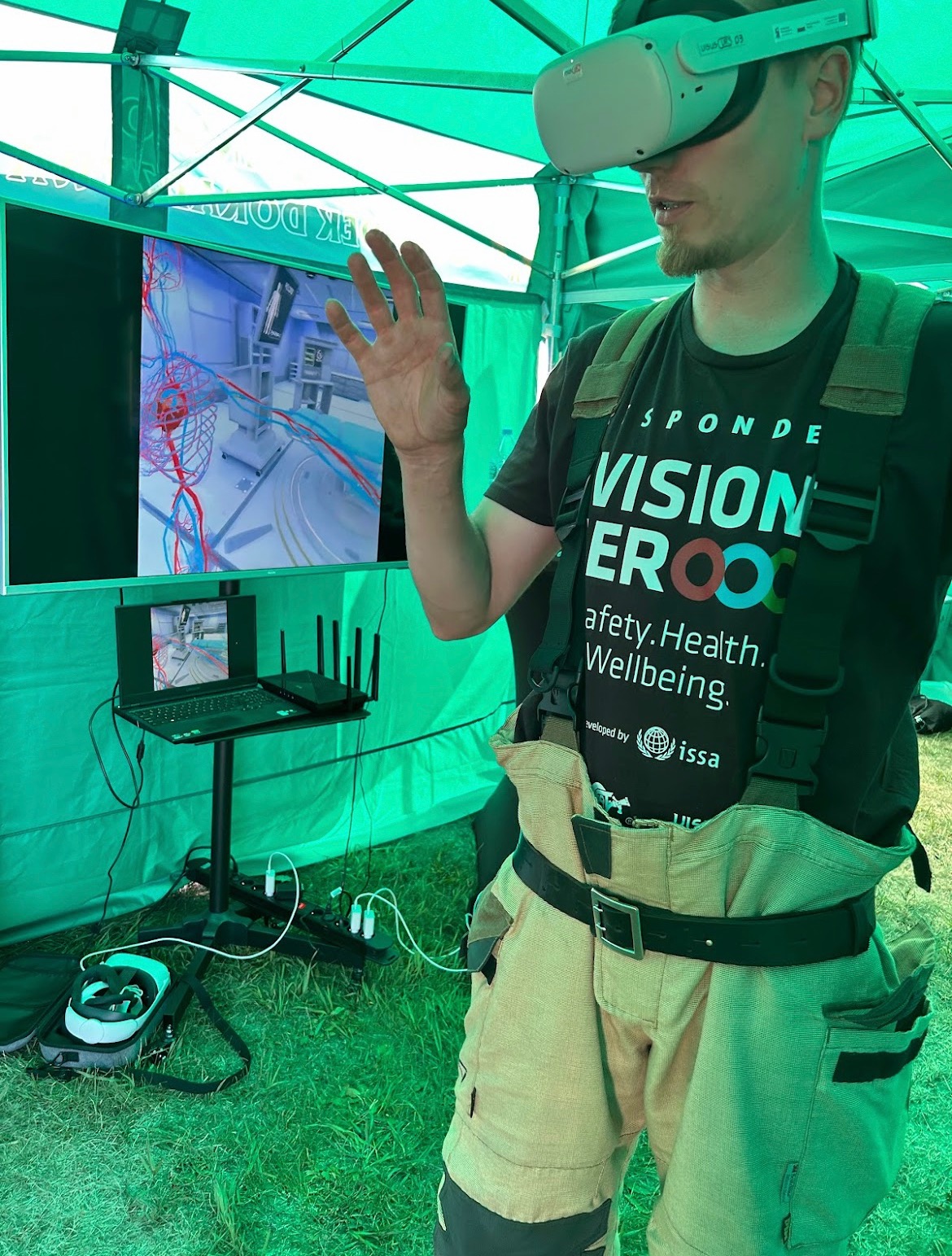 Mężczyzna w kombinezonie roboczym i goglach VR, gestykuluje przed ekranem prezentującym wirtualną symulację wnętrza, prawdopodobnie w kontekście szkolenia z bezpieczeństwa.