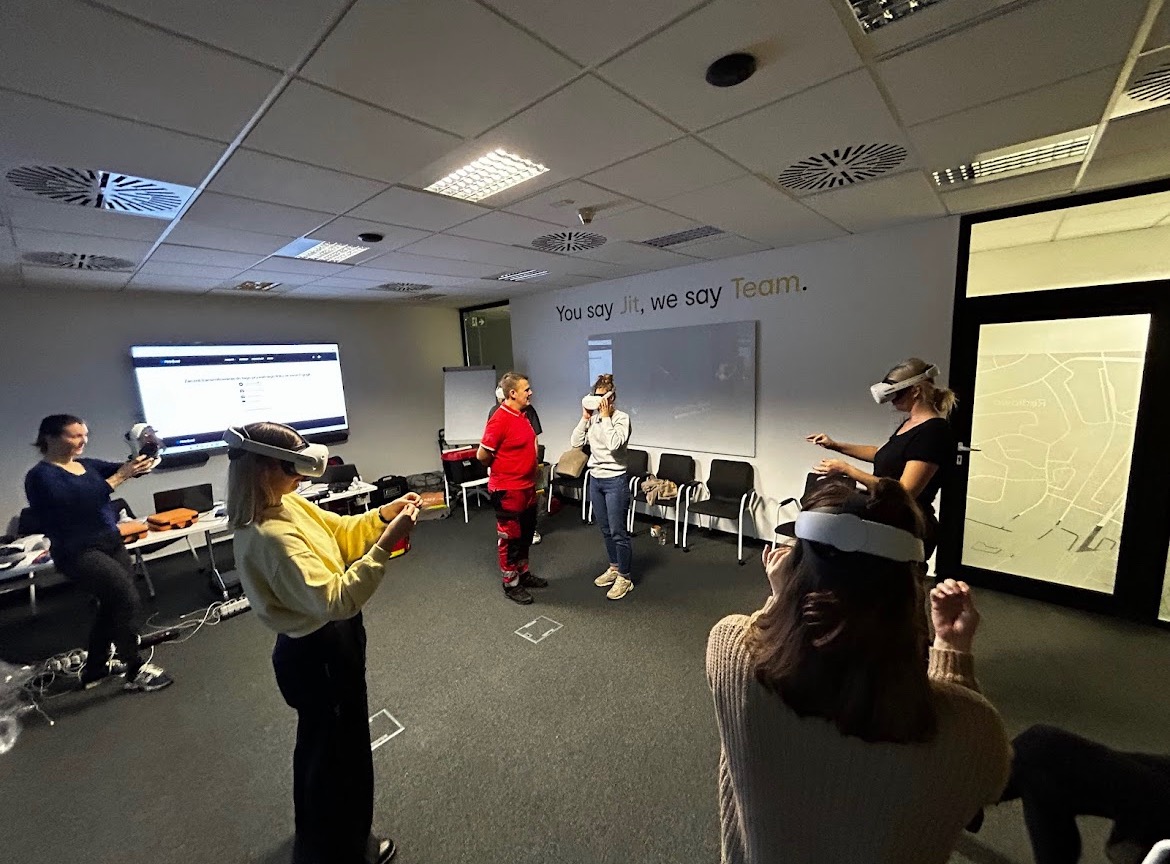 Grupa osób w sali szkoleniowej z okularami VR uczestniczy w symulacji pierwszej pomocy; instruktor w czerwonym kombinezonie obserwuje uczestników.