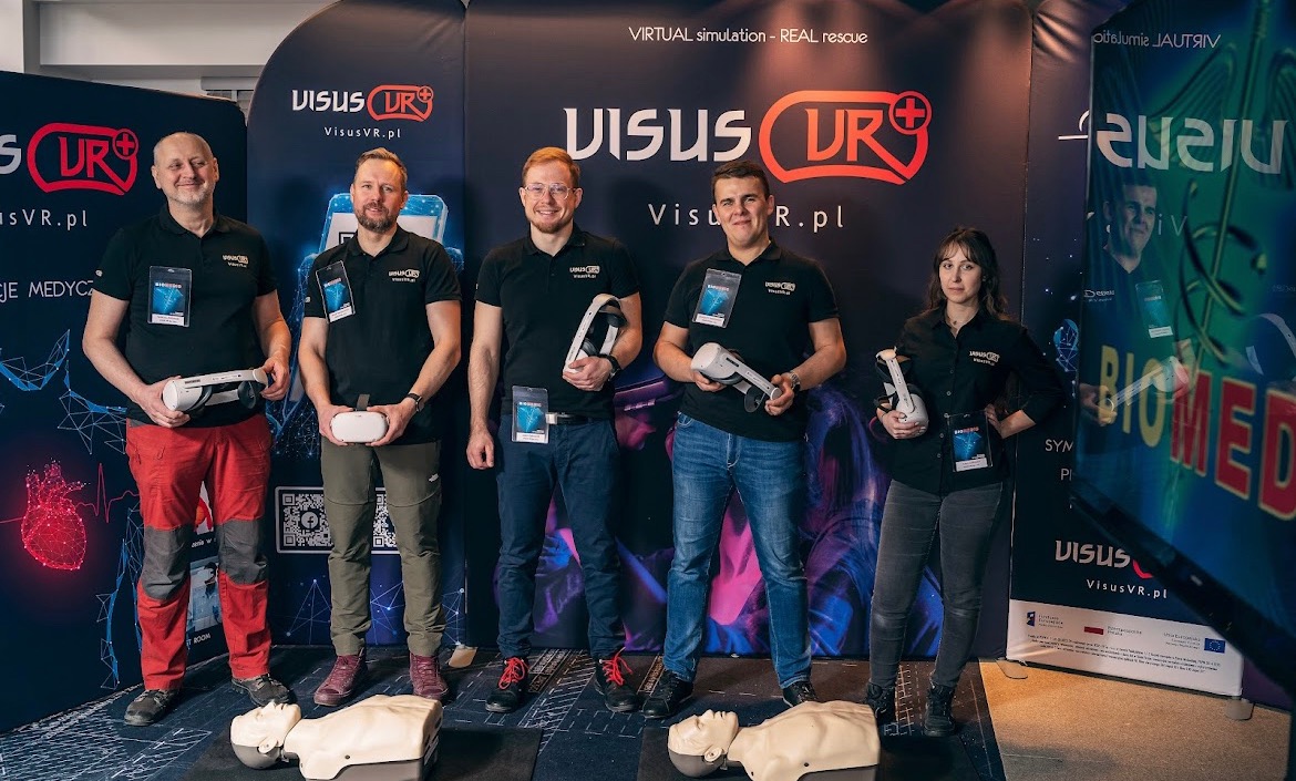 Grupa pięciu osób prezentuje symulację pierwszej pomocy z wykorzystaniem wirtualnej rzeczywistości, trzymając zestawy VR na tle stoiska z logo firmy Visus VR i manekinami do ćwiczeń resuscytacji.
