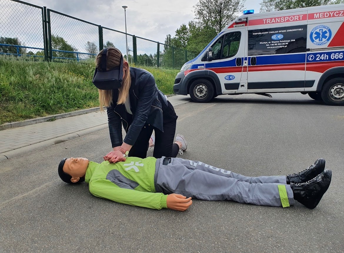 Osoba w goglach VR ćwiczy resuscytację krążeniowo-oddechową na manekinie na asfalcie, w tle ambulans.