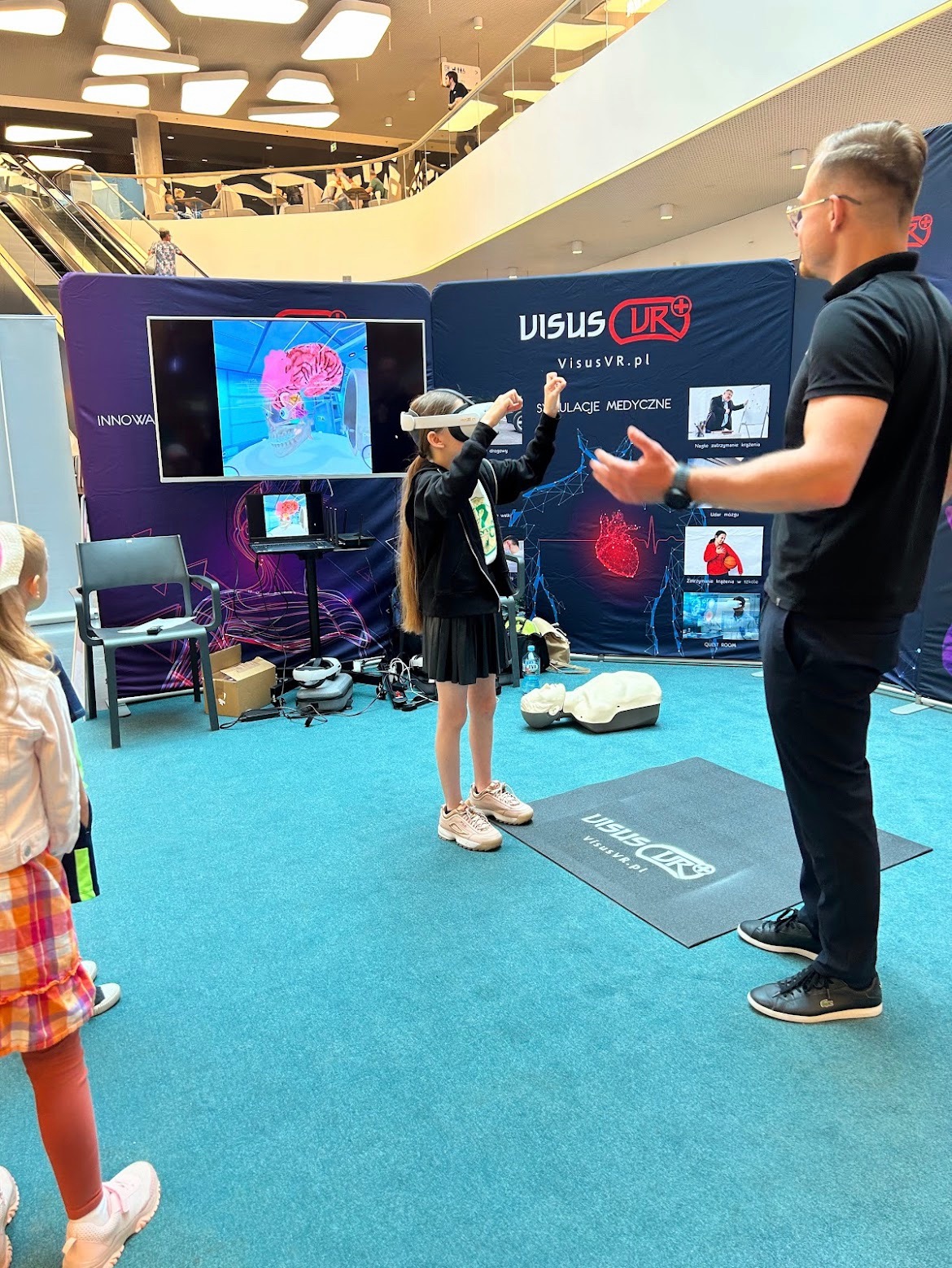 Dziewczynka w okularach VR symuluje czynności medyczne na tle stoiska VisusVR.pl, obok stoi instruktor, w tle manekin do resuscytacji i ekran prezentujący wnętrze ludzkiego mózgu.