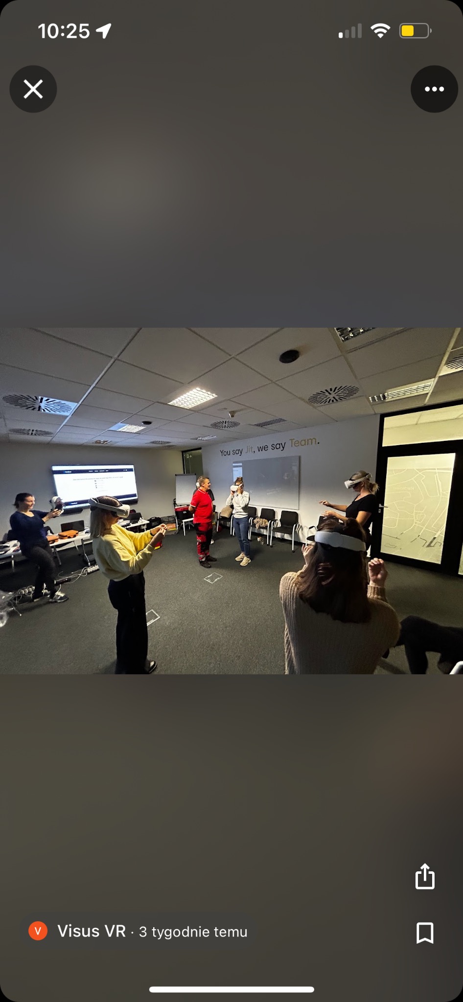 Grupa osób w okularach VR uczestniczy w symulacji w sali szkoleniowej, z widocznym monitorem i napisem 'You say Jit, we say Team.' na ścianie.
