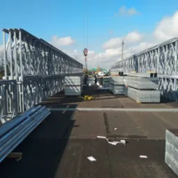 Montaż elementów konstrukcyjnych domu modułowego na dachu, widoczne stalowe ramy kratownicowe, palety z panelami i żuraw budowlany w tle.