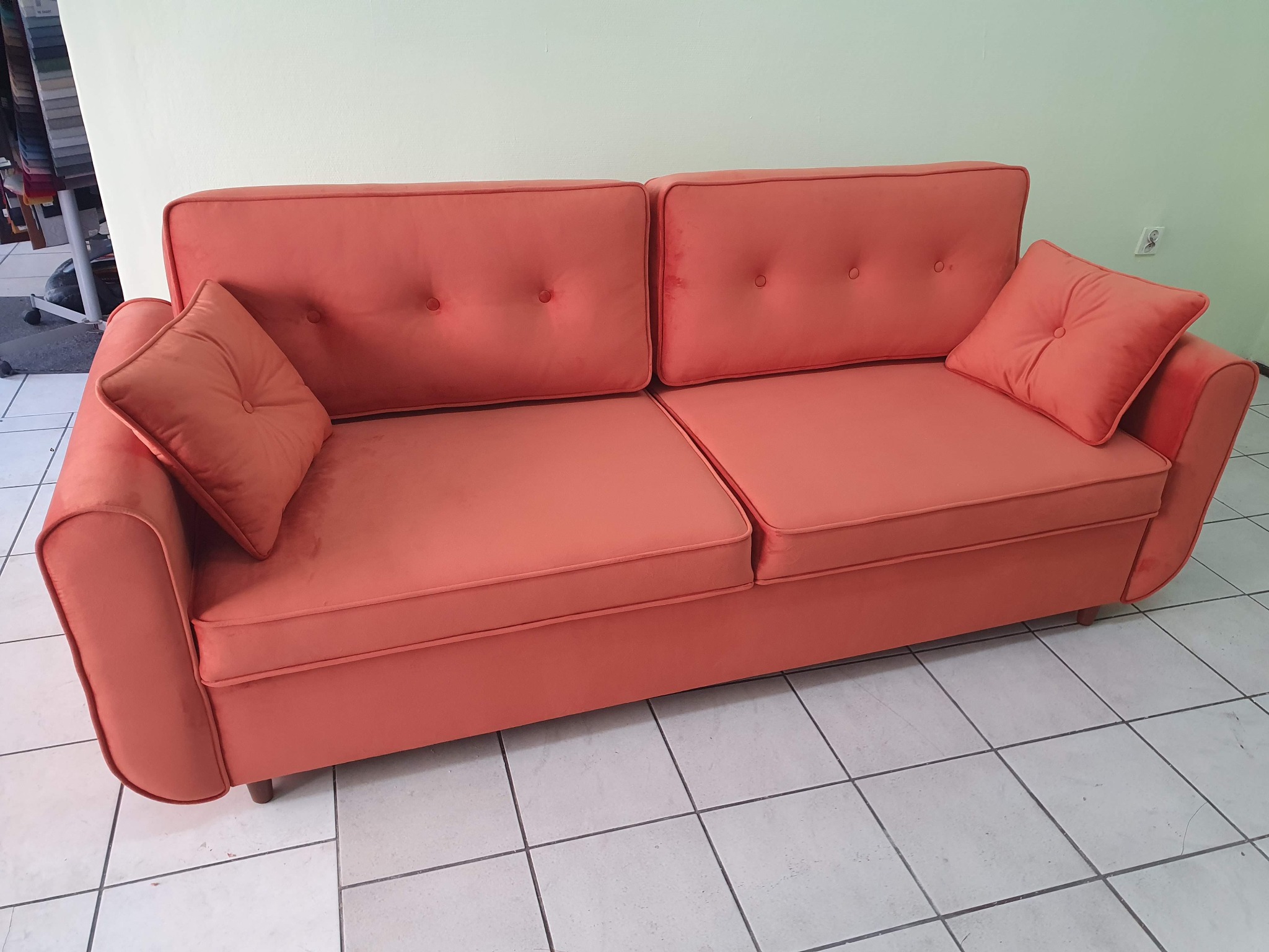 Koralowa sofa dwuosobowa z poduszkami, tapicerowana materiałem z guzikami, na drewnianych nóżkach, ustawiona na białej podłodze z płytek.