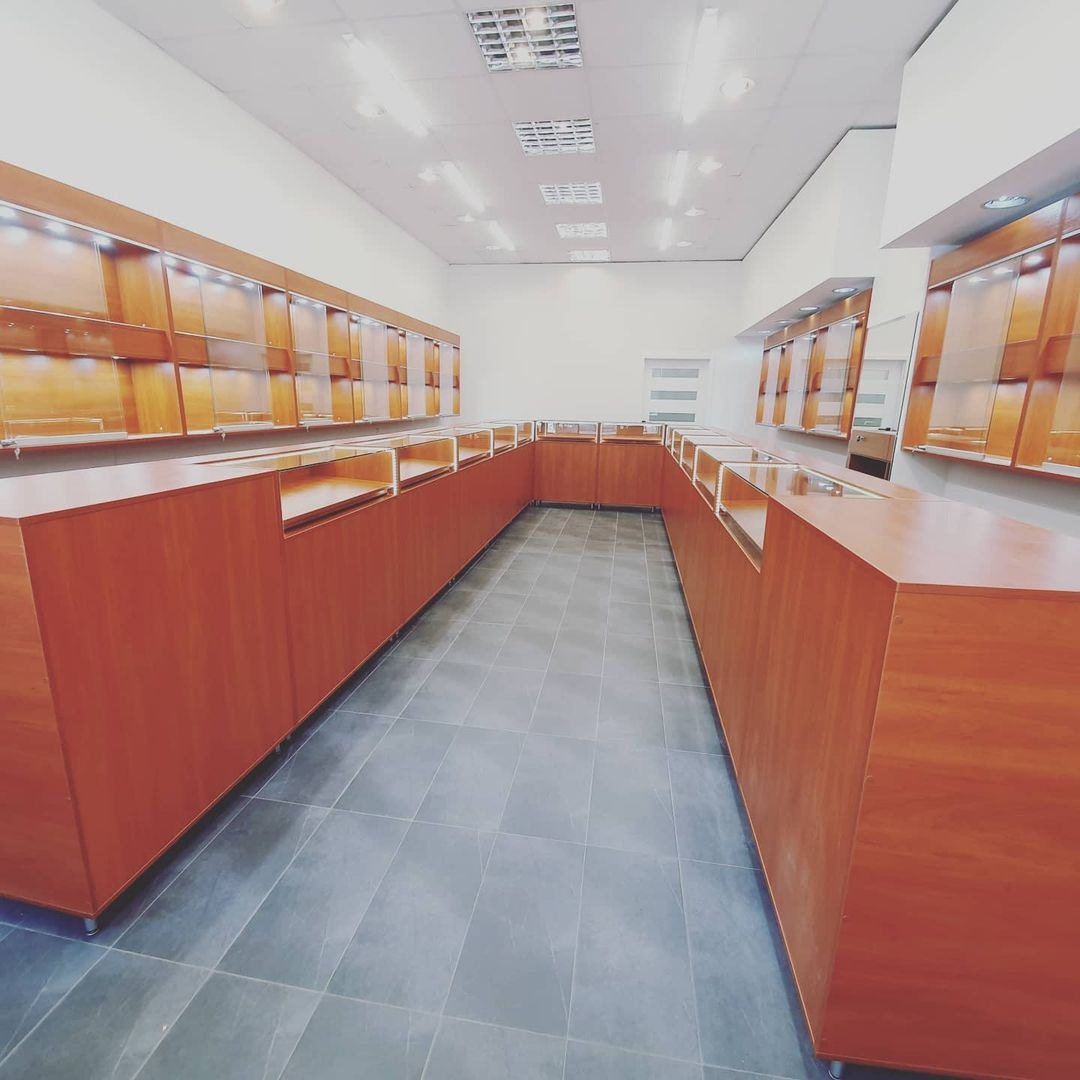 Wnętrze sklepu z drewnianymi regałami i przeszklonymi gablotami wzdłuż ścian, podświetlane półki, szara podłoga z płytek.