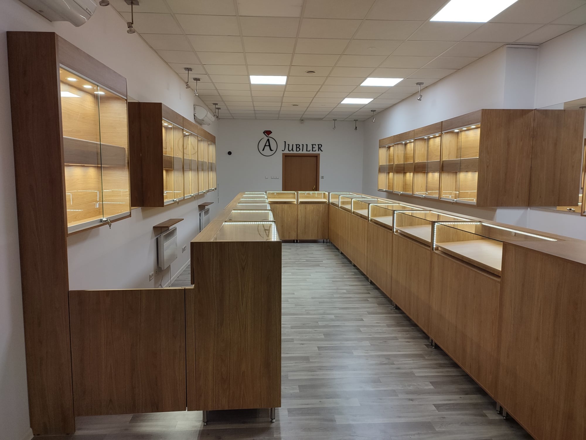 Wnętrze sklepu jubilerskiego z drewnianymi witrynami ekspozycyjnymi podświetlonymi LED, widok na logo firmy na ścianie.