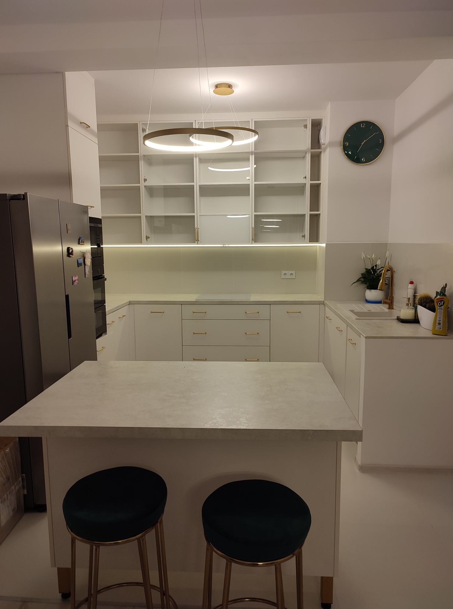 Nowoczesna, jasna kuchnia z wyspą, białymi szafkami z delikatnymi złotymi uchwytami, okrągłym złotym oświetleniem i ciemnozielonym zegarem na ścianie.