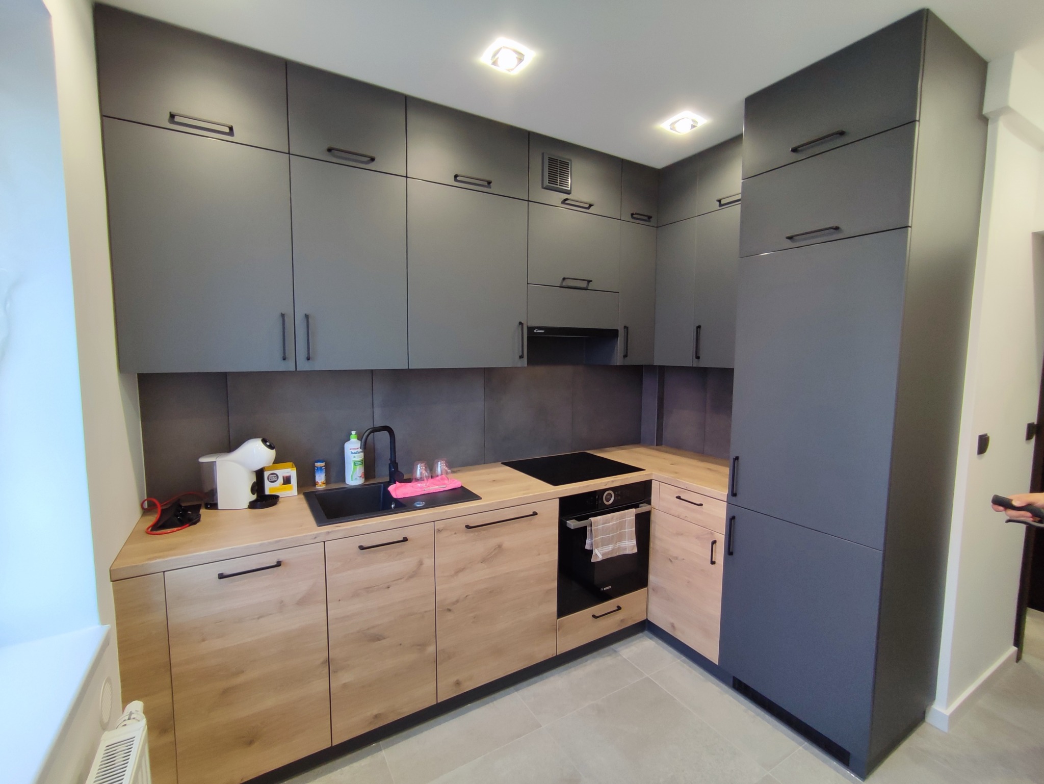 Nowoczesna kuchnia z szarymi szafkami górnymi i drewnopodobnymi dolnymi, czarny zlew i płyta indukcyjna, oświetlenie punktowe, ekspres do kawy na blacie.