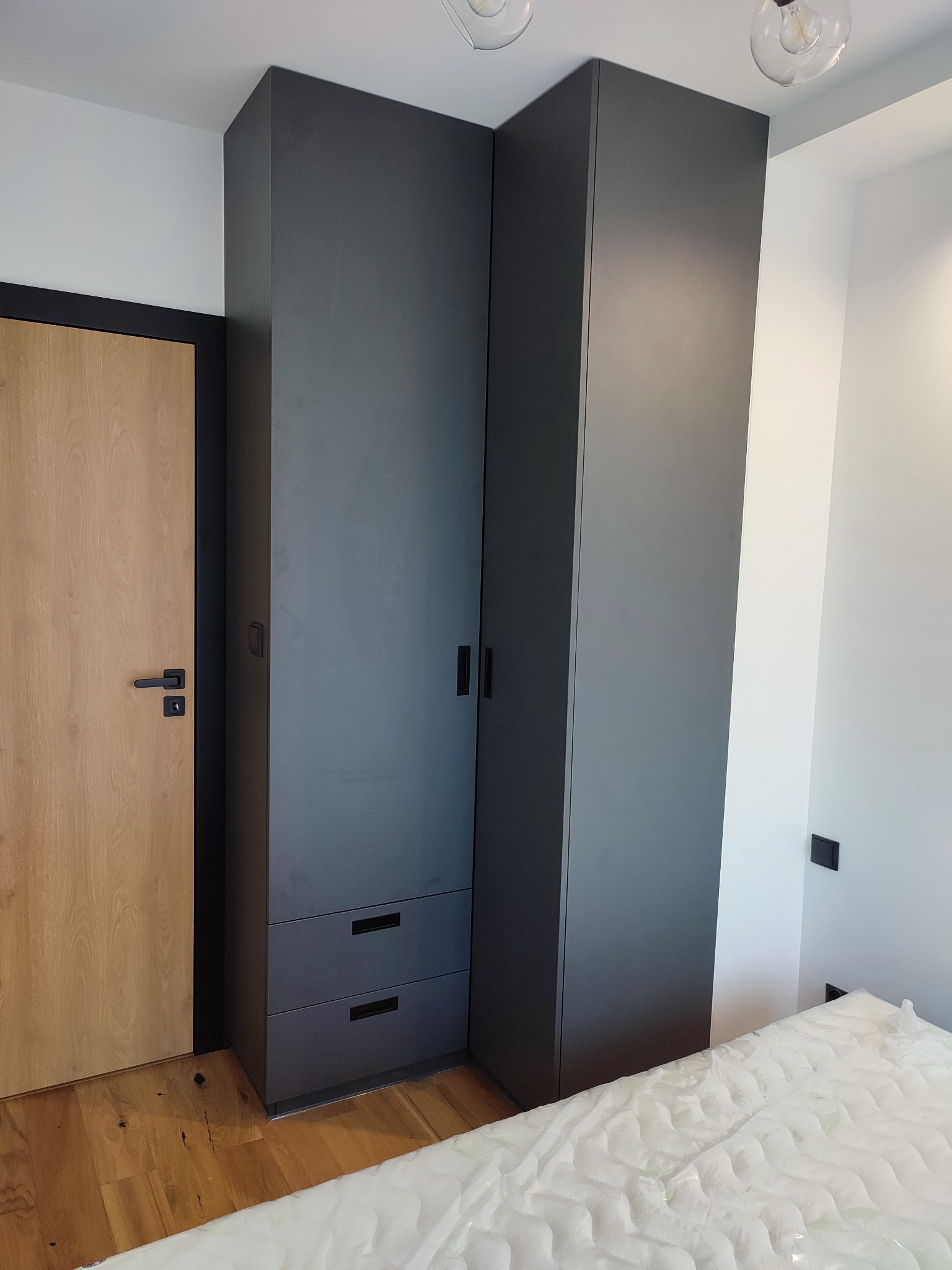 Ciemnoszara szafa wnękowa z szufladami, zintegrowana z drewnianą podłogą i drzwiami w nowoczesnym wnętrzu, widok na fragment materaca.