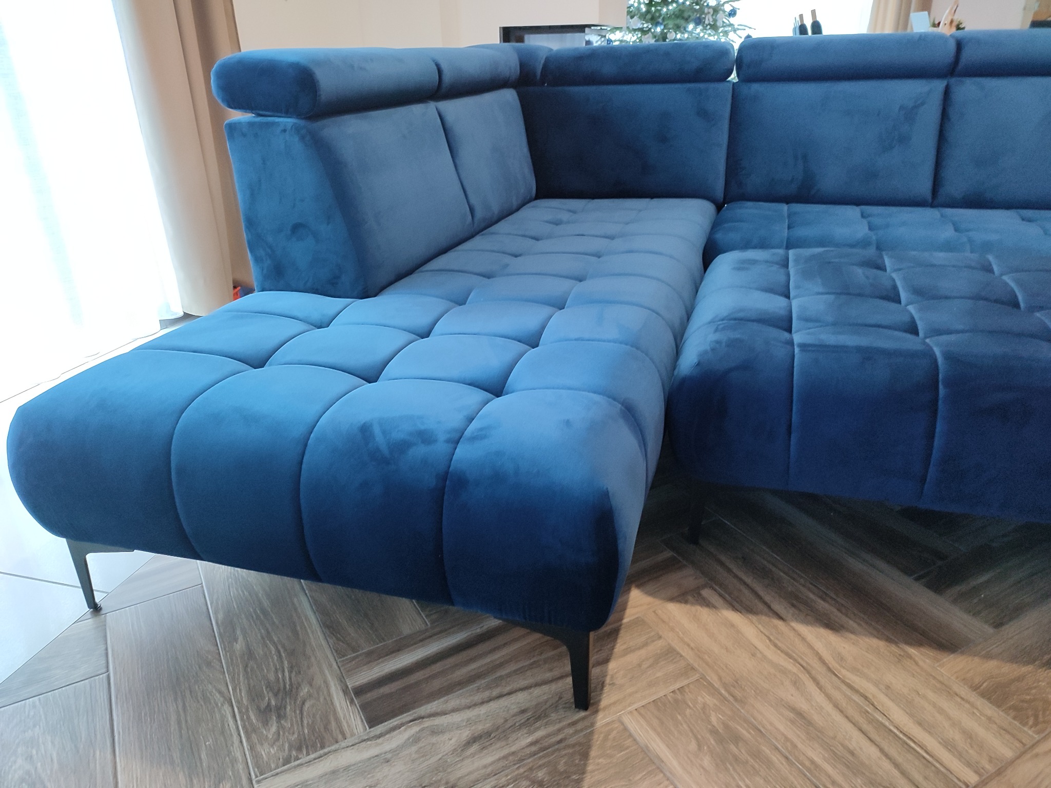 Narożnik modułowy obity niebieskim welurem, z regulowanymi zagłówkami i pikowanym siedziskiem, na tle parkietu w jodełkę.