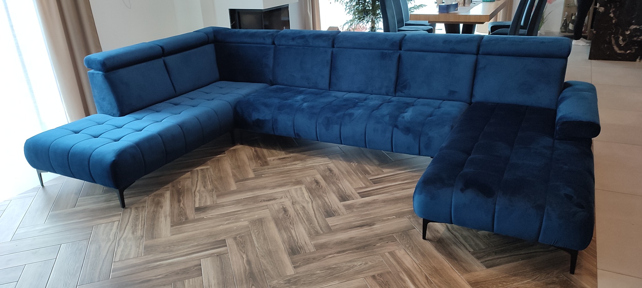 Narożnik tapicerowany w odcieniach granatu, z pikowanymi siedziskami i oparciami, ustawiony na podłodze z wzorem jodełki.