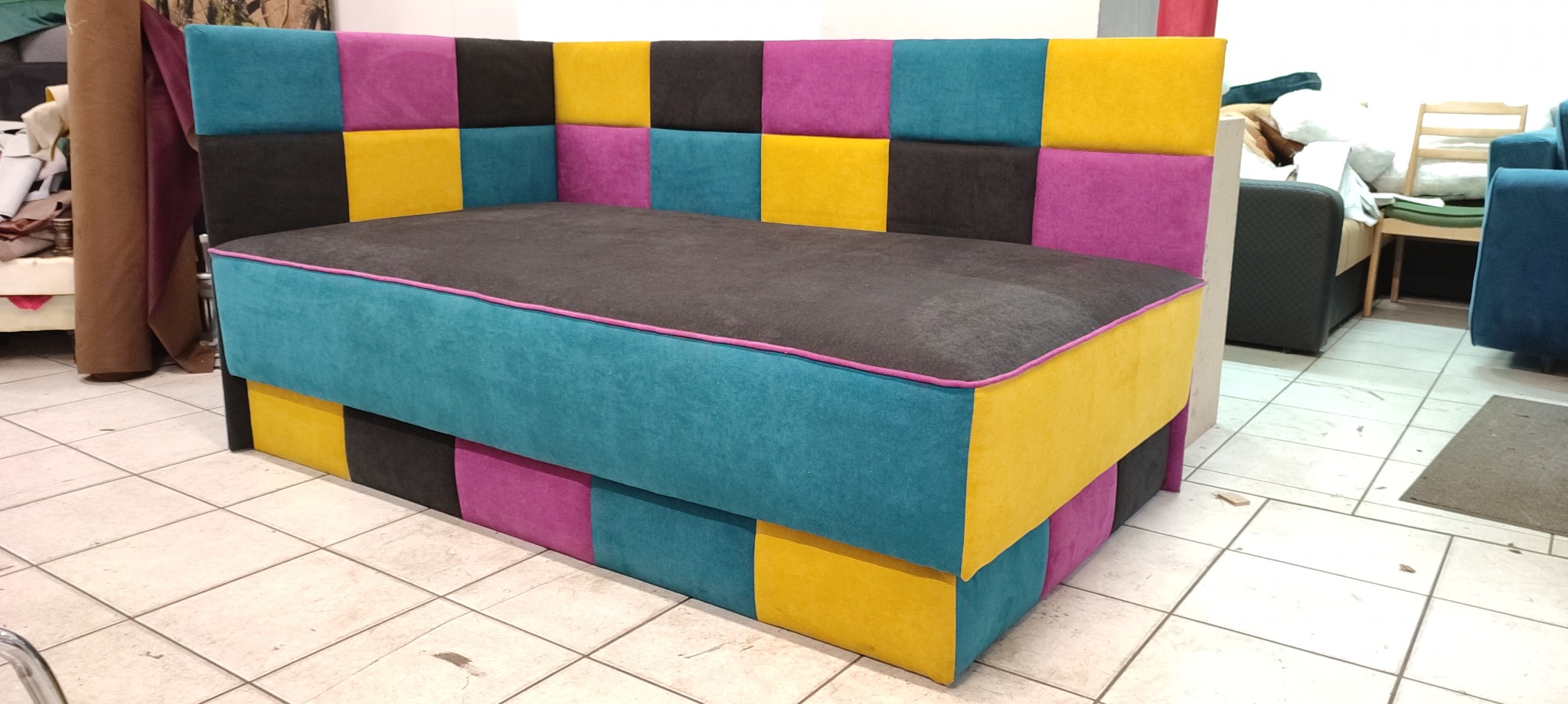 Tapicerowany narożnik z patchworkowym wzorem w odcieniach turkusu, fioletu, żółci i czerni, wykończony różową lamówką, widoczny w warsztacie tapicerskim.