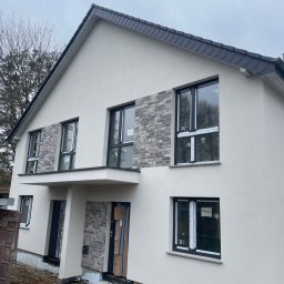 Hlodovyu - Nowoczesny dom jednorodzinny w Olsztynie z ocieploną białą elewacją i fragmentami dekoracyjnej okładziny imitującej szarą cegłę, widoczne okna z czarnymi ramami i przygotowane wejście.