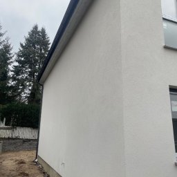 Hlodovyu - Fragment bocznej ściany budynku z jasną, świeżo nałożoną elewacją o strukturze baranka, widoczne okno z folią ochronną, w tle drzewa iglaste i fragment ogrodzenia.