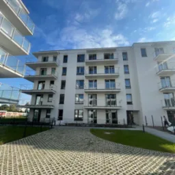 Nowoczesny, biały budynek wielorodzinny z balkonami o szklanych balustradach, widok z dołu na tle błękitnego nieba z chmurami, z przodu ażurowy parking z zielenią.