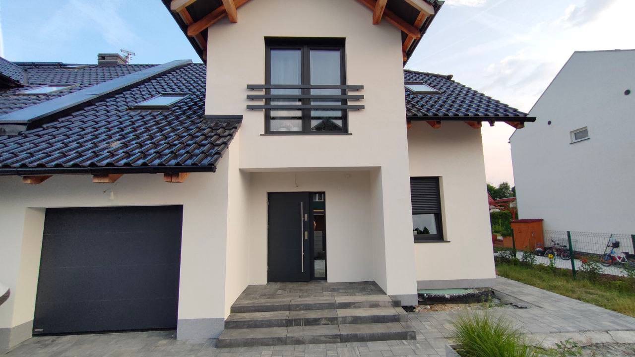 Nowoczesny dom jednorodzinny z jasną elewacją, ciemną stolarką okienną i drzwiową, oraz garażem z bramą segmentową w kolorze antracytowym.