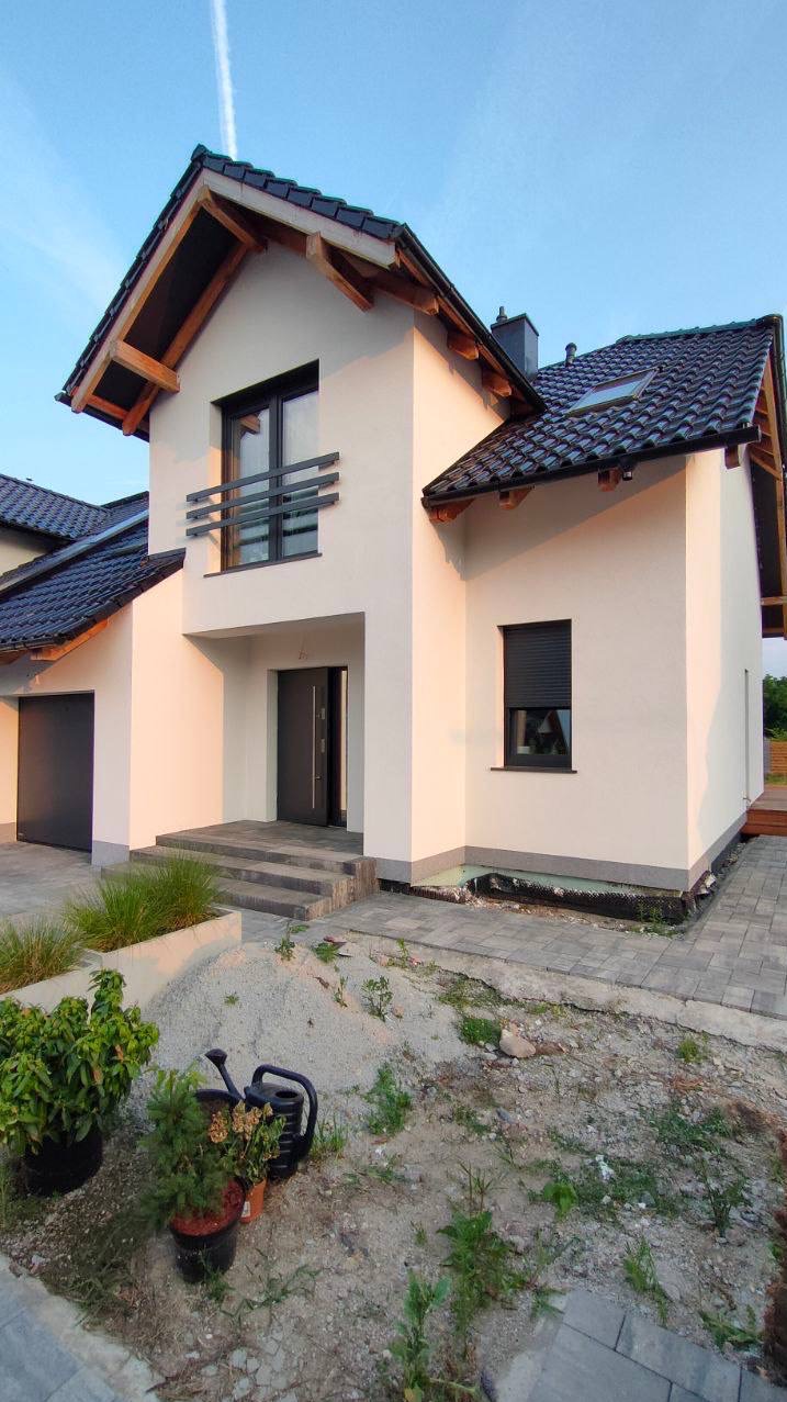 Nowoczesny dom jednorodzinny z jasną elewacją, ciemną stolarką okienną i dachówką, widoczne prace wykończeniowe wokół budynku, w tym ułożona kostka brukowa i rośliny w donicach.