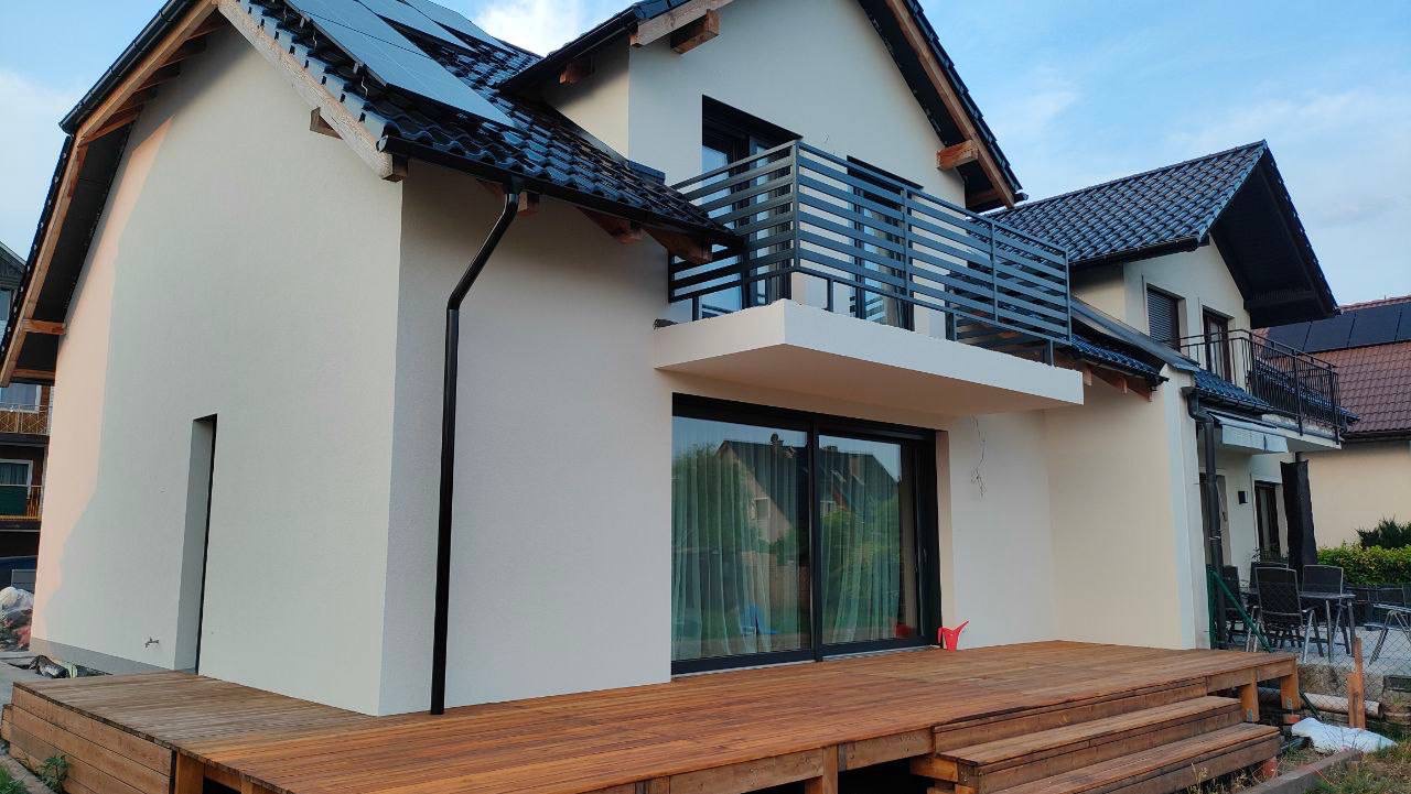 Nowoczesny dom z jasną elewacją, ciemną dachówką i drewnianym tarasem, widok z boku na balkon z metalową balustradą.