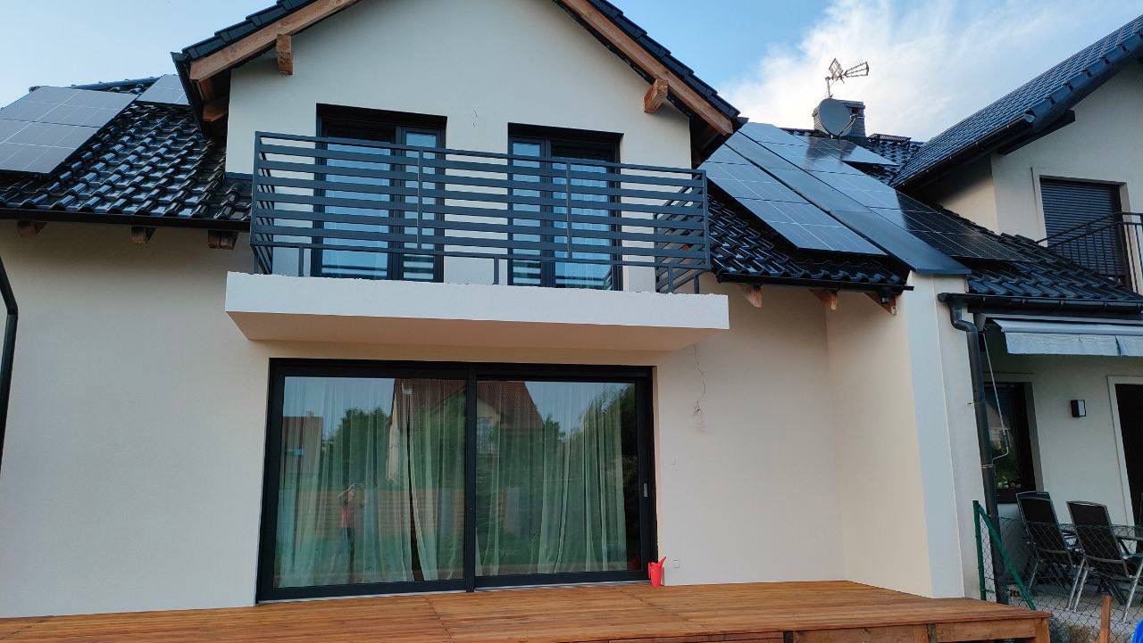 Dwukondygnacyjny dom z jasną elewacją, balkonem z czarną, poziomą balustradą i panelami słonecznymi na dachu, widoczny z zewnątrz.