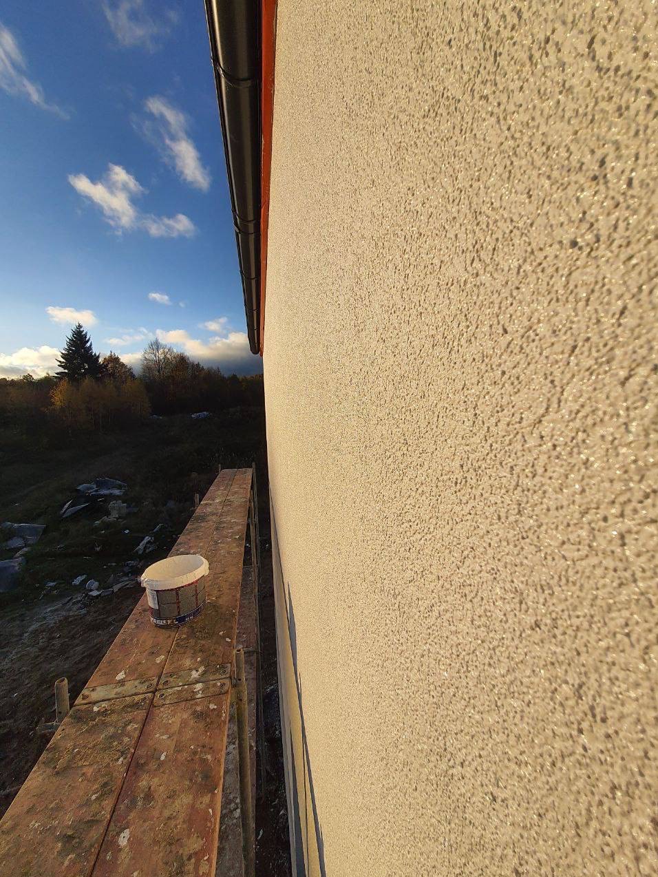 Świeżo otynkowana elewacja budynku w kolorze beżowym, widoczna z rusztowania, z wiadrem farby na pierwszym planie i fragmentem nieba w tle.