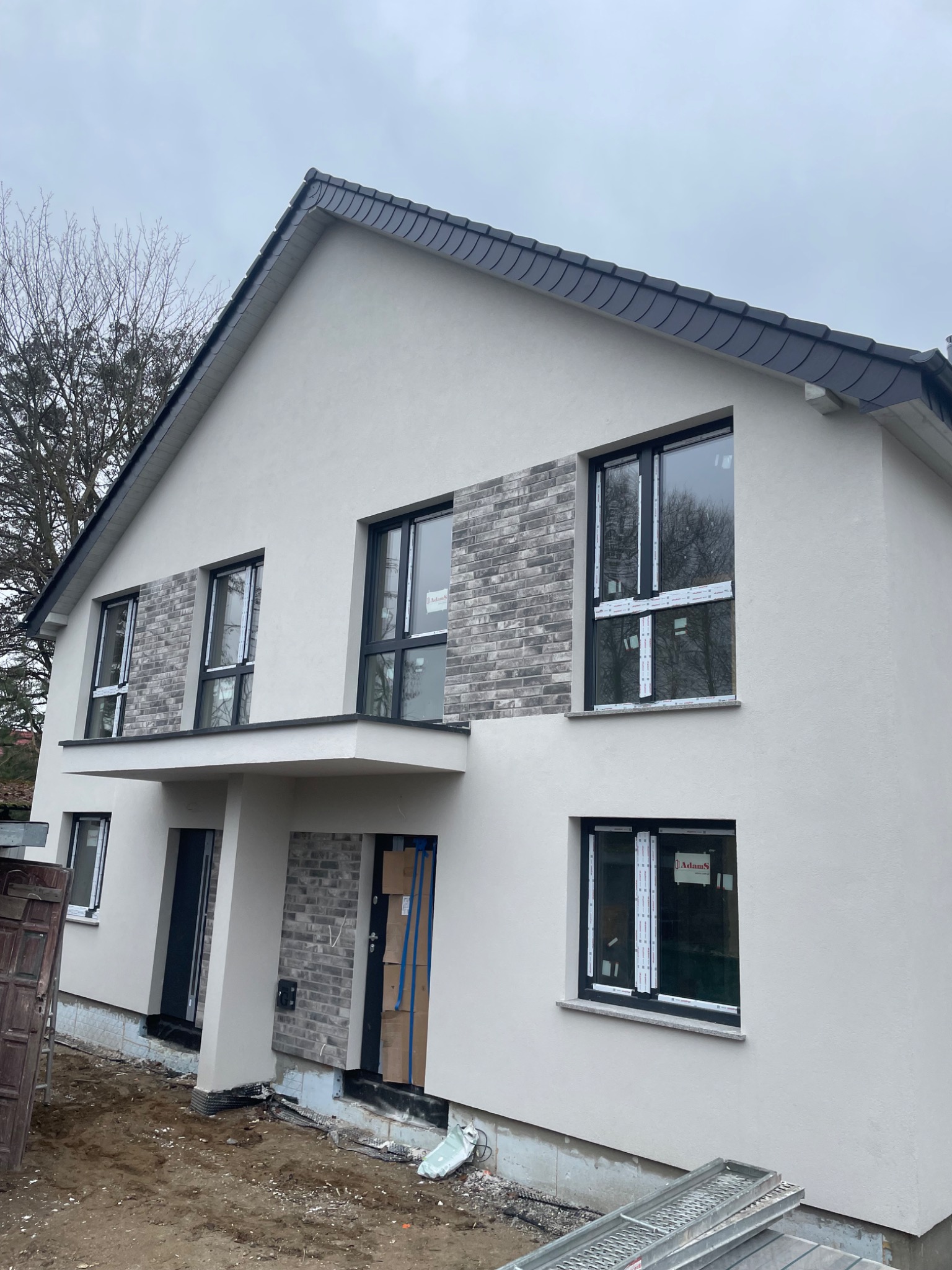 Nowoczesny dom jednorodzinny w Olsztynie z ocieploną białą elewacją i fragmentami dekoracyjnej okładziny imitującej szarą cegłę, widoczne okna z czarnymi ramami i przygotowane wejście.