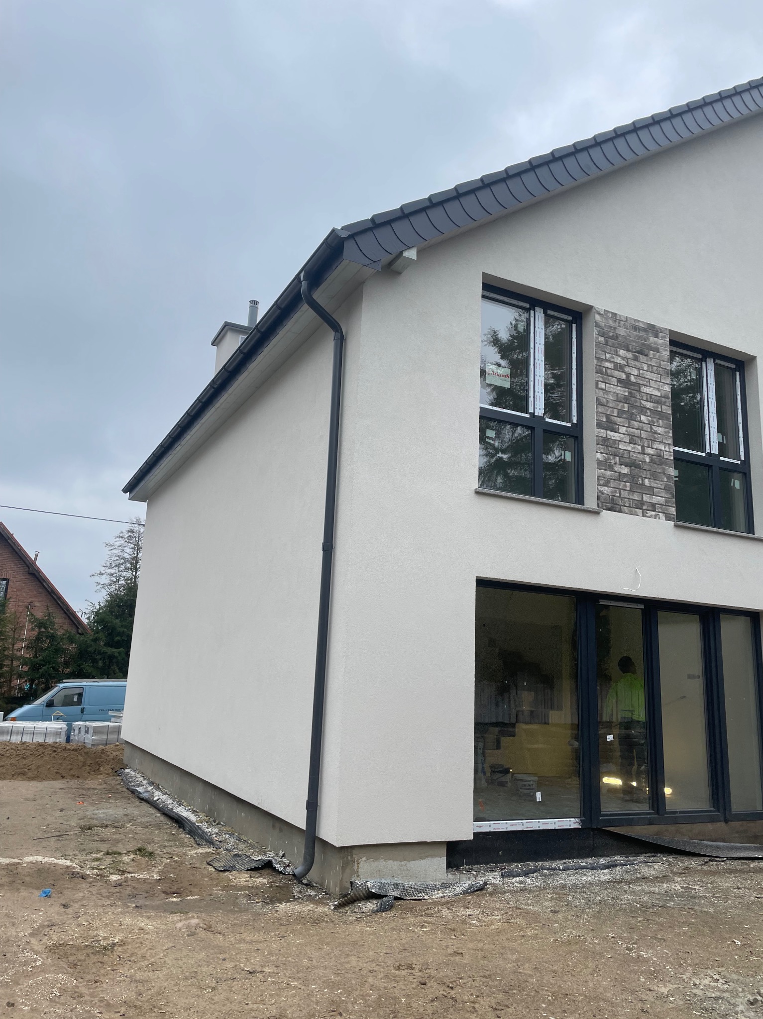 Fragment nowo wybudowanego domu z jasną elewacją, czarnymi oknami i wykończeniem z szarej cegły klinkierowej na fragmentach ściany; widoczne rynny, dachówka i teren wokół budynku w trakcie prac...