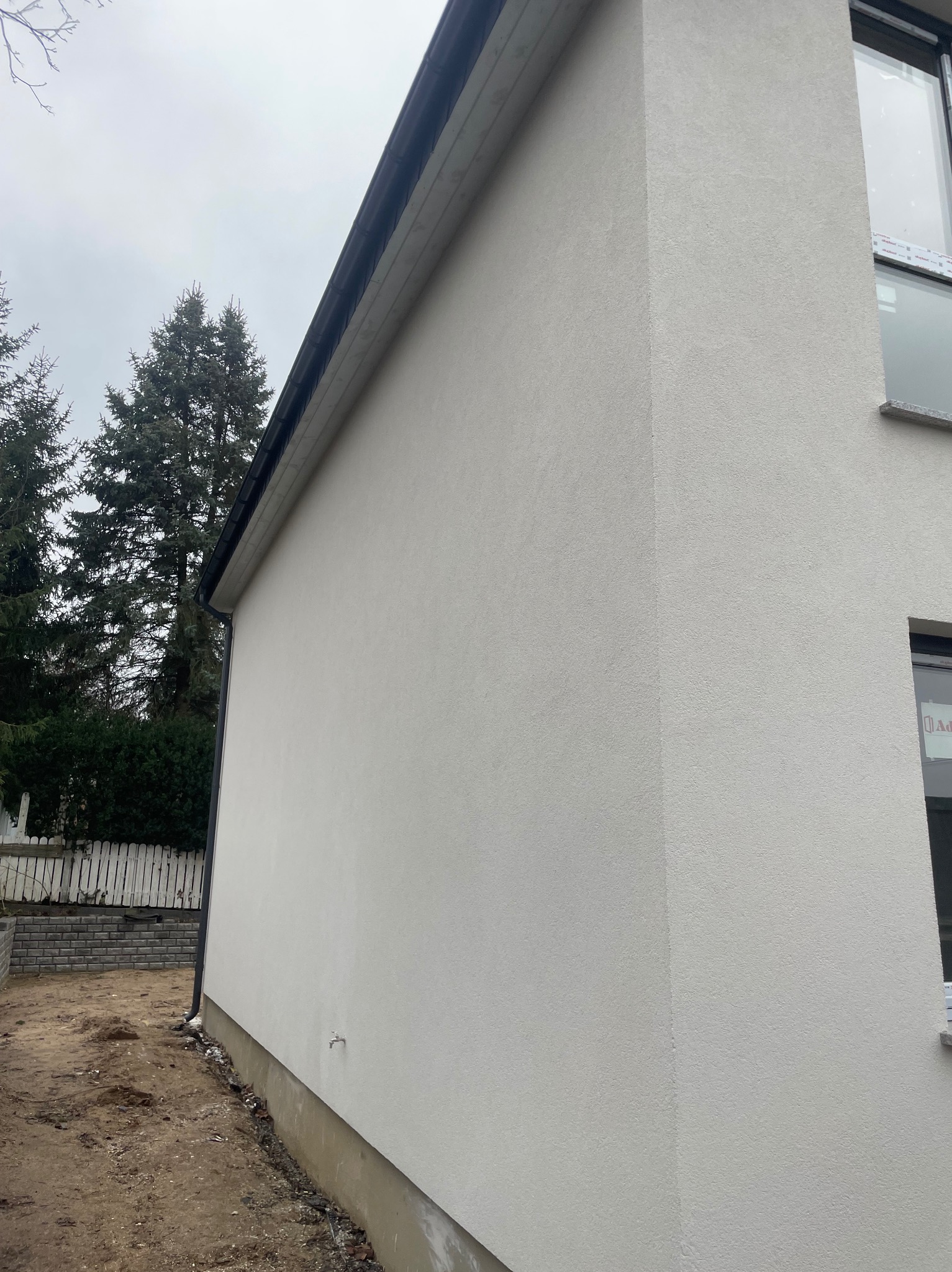 Fragment bocznej ściany budynku z jasną, świeżo nałożoną elewacją o strukturze baranka, widoczne okno z folią ochronną, w tle drzewa iglaste i fragment ogrodzenia.