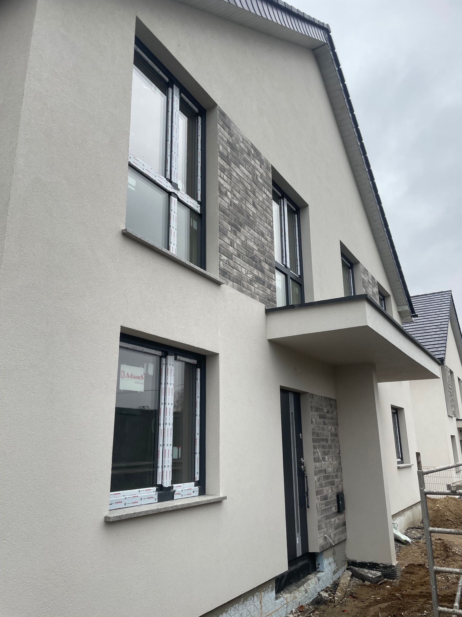 Nowoczesny dom w trakcie wykańczania elewacji: jasnoszary tynk strukturalny kontrastuje z fragmentami ściany wyłożonymi szarą cegłą, czarne ramy okienne z folią ochronną.