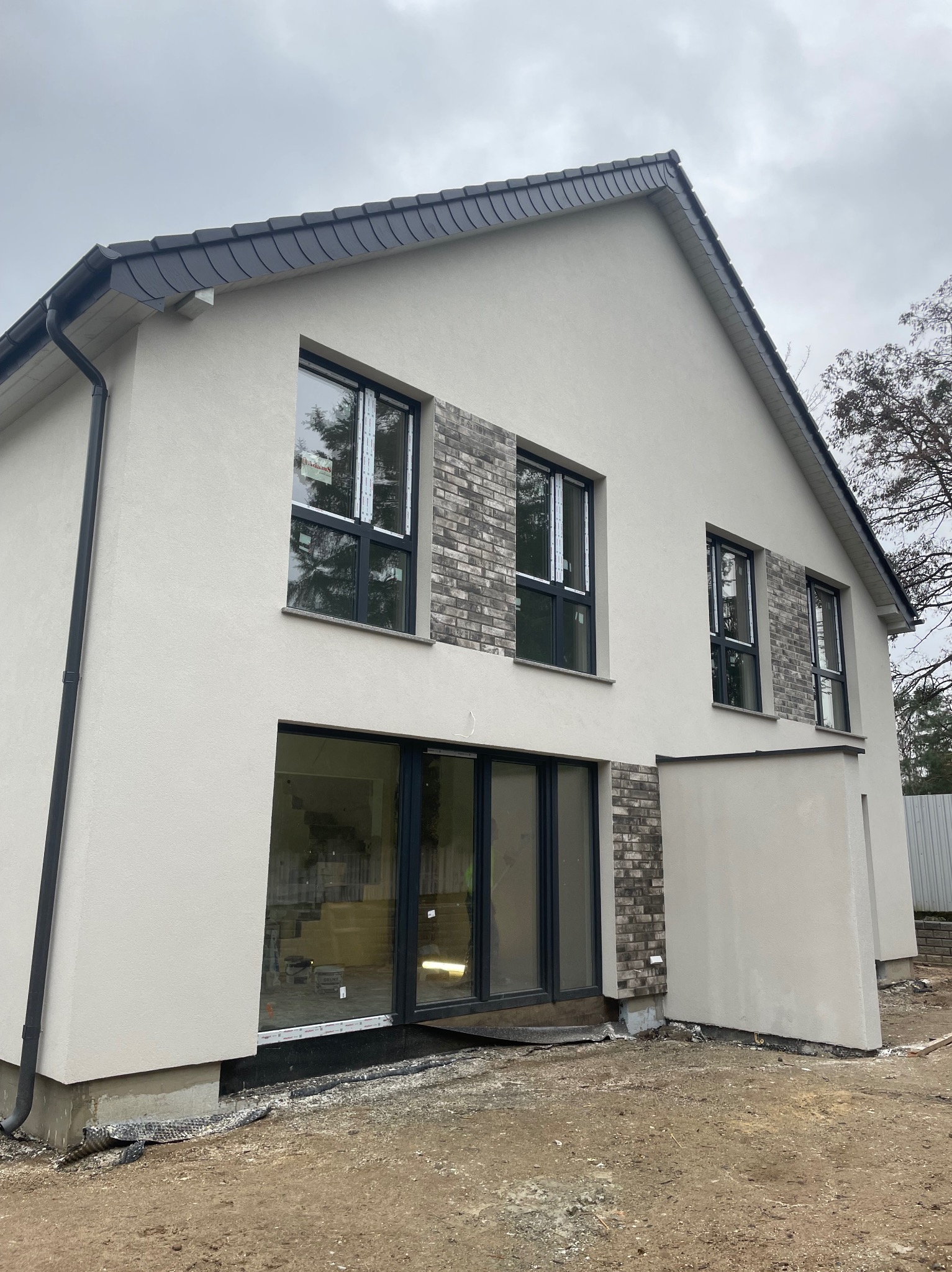 Dwukondygnacyjny dom jednorodzinny z jasną elewacją, fragmentami wykończonymi szarą cegłą, czarnymi ramami okiennymi i drzwiami przesuwnymi, widoczne prace wykończeniowe wokół budynku.