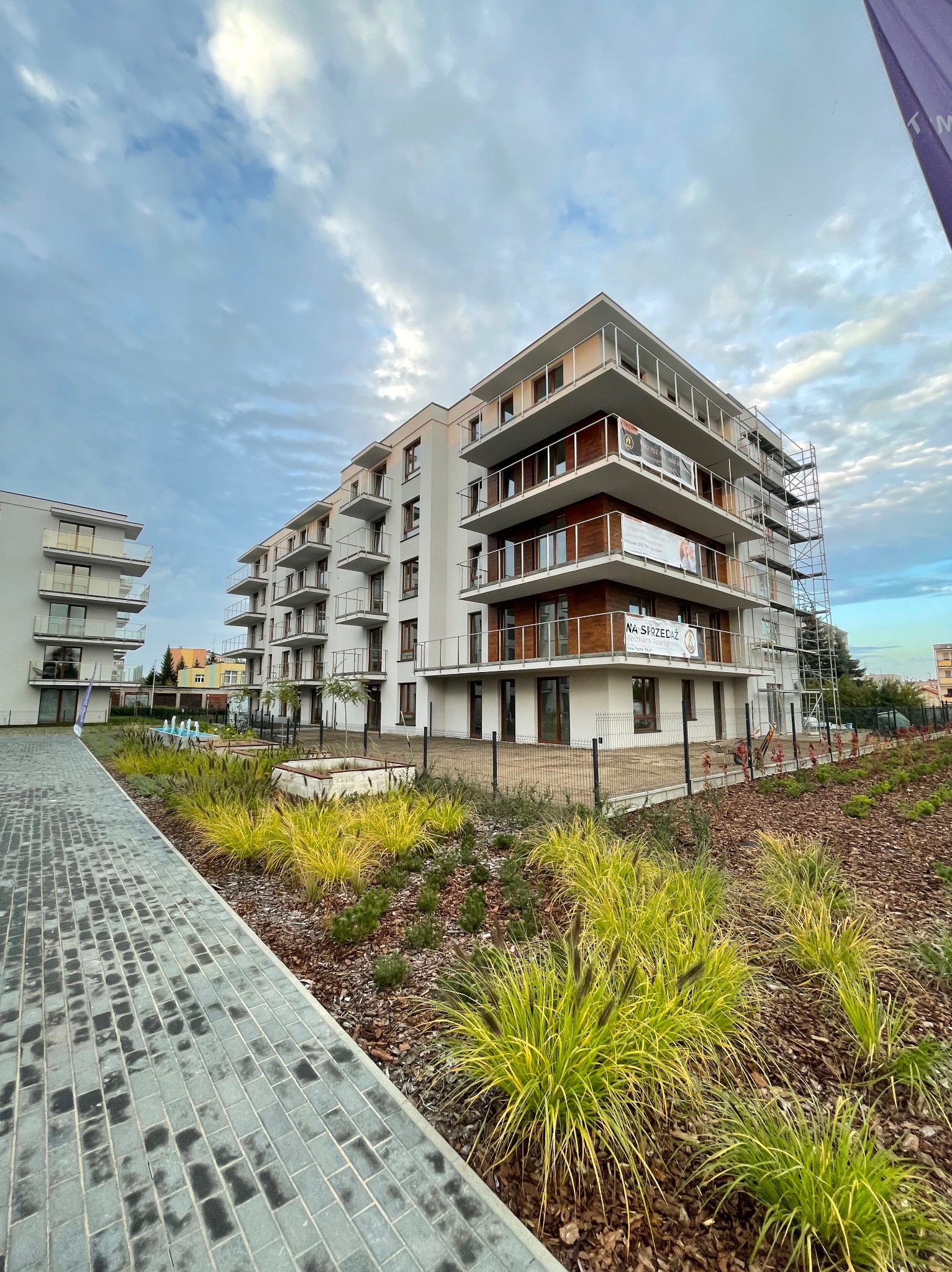 Nowoczesny budynek apartamentowy z balkonami, w trakcie wykańczania elewacji, z widocznymi rusztowaniami i banerami reklamowymi 'Na Sprzedaż', otoczony zagospodarowaną zielenią z fontanną...
