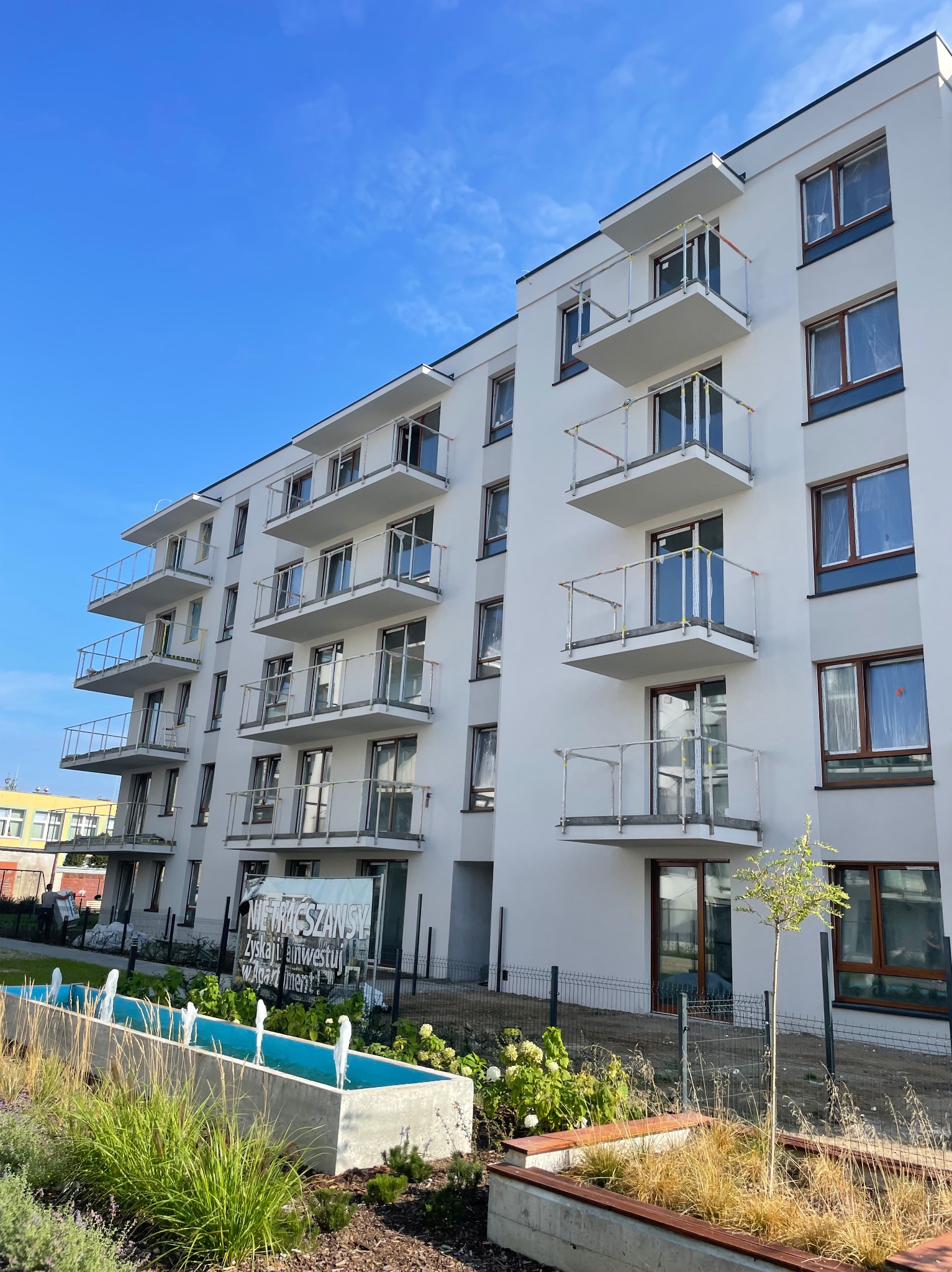 Nowoczesny budynek wielorodzinny z białymi elewacjami i balkonami z przeszklonymi balustradami, w tle błękitne niebo i fontanna przed budynkiem.